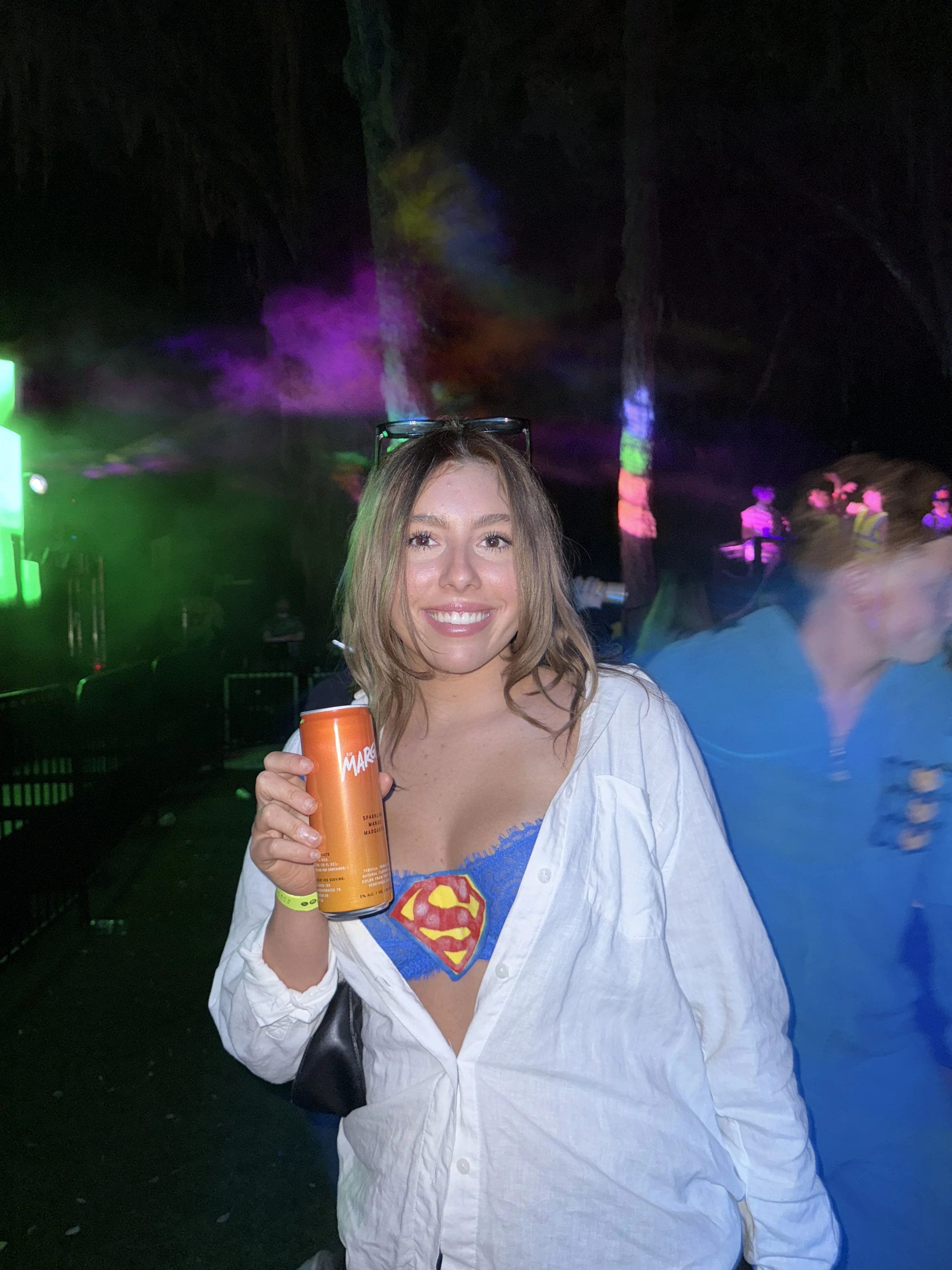 A young woman with shoulder-length brown hair smiling and holding an orange can at a nighttime event with colorful lights and a dark background.