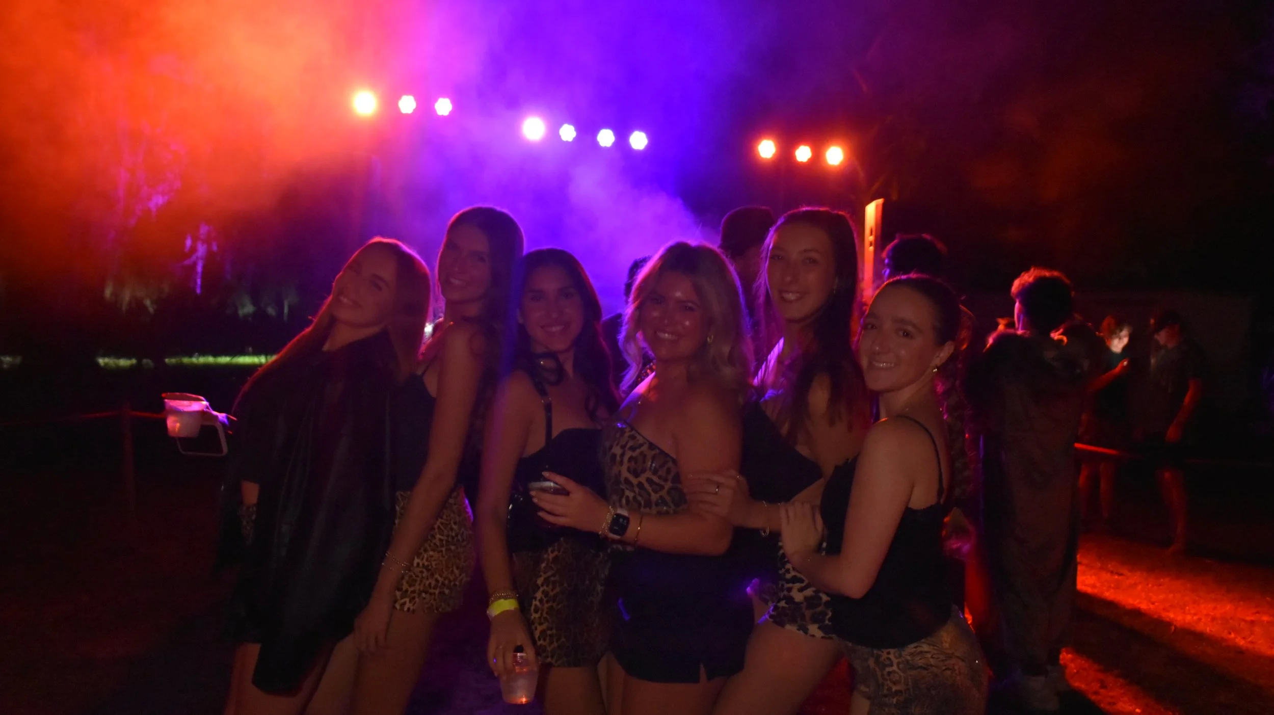 Group of women at a nighttime outdoor music or dance event with colorful stage lights, some wearing animal print clothing.