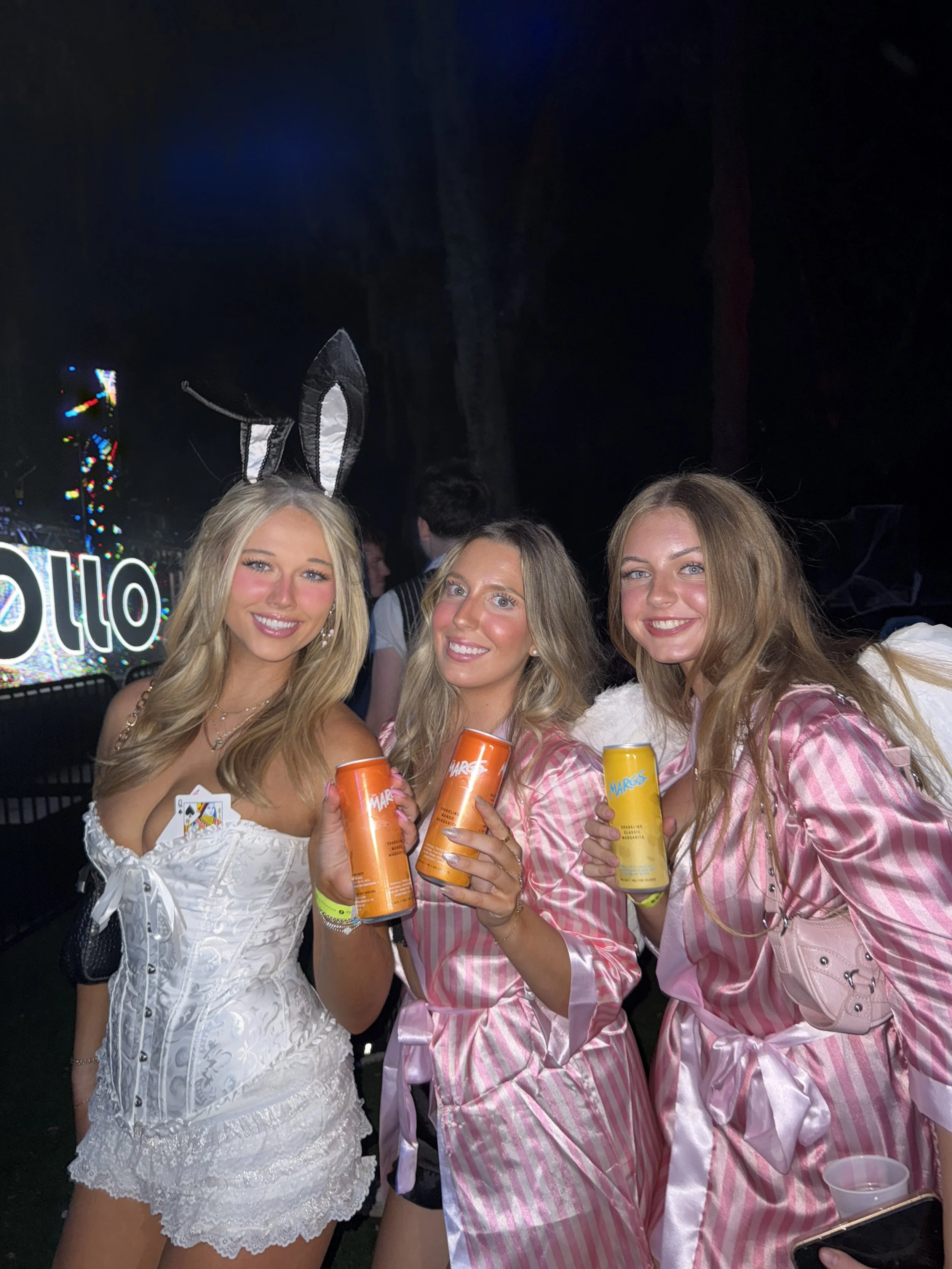 Three young women dressed in pink and white striped pajamas and a white corset with lace, holding cans of beverages, at a nighttime event with a dark background and colorful lights.