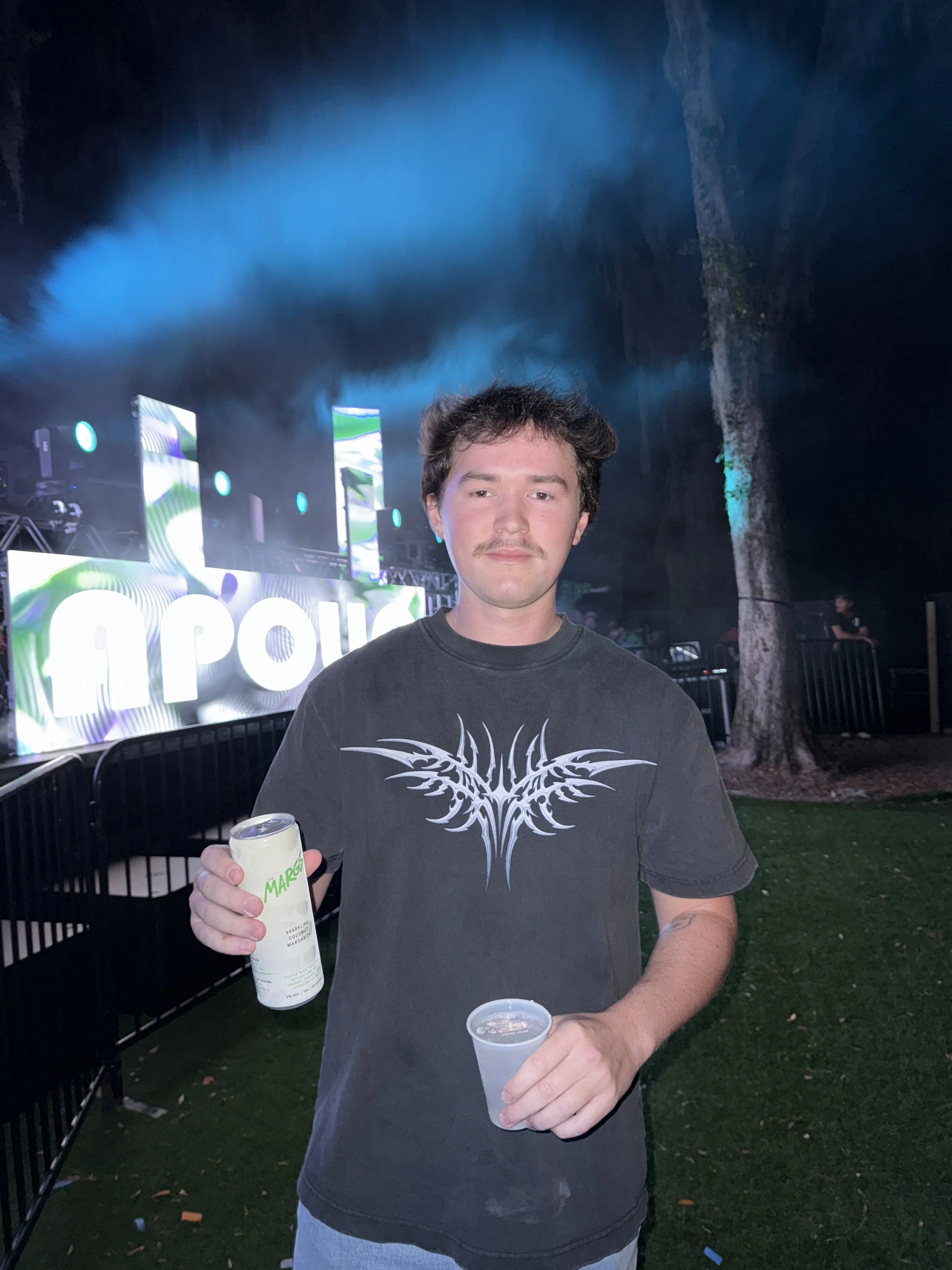 A young man with a mustache and dark, wavy hair standing outdoors at night, holding a drink can and a cup, with a stage and colorful lights in the background.