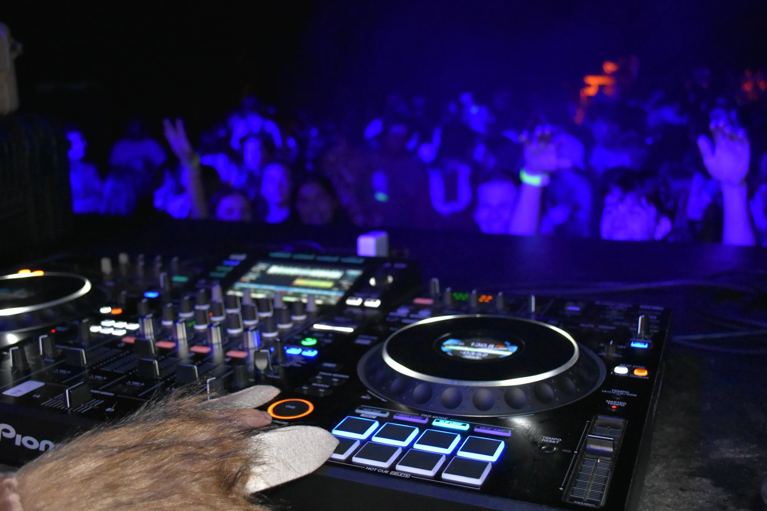 DJ's hand on a Pioneer DJ controller at a nightclub with a crowd dancing in the background, illuminated by colorful lights.