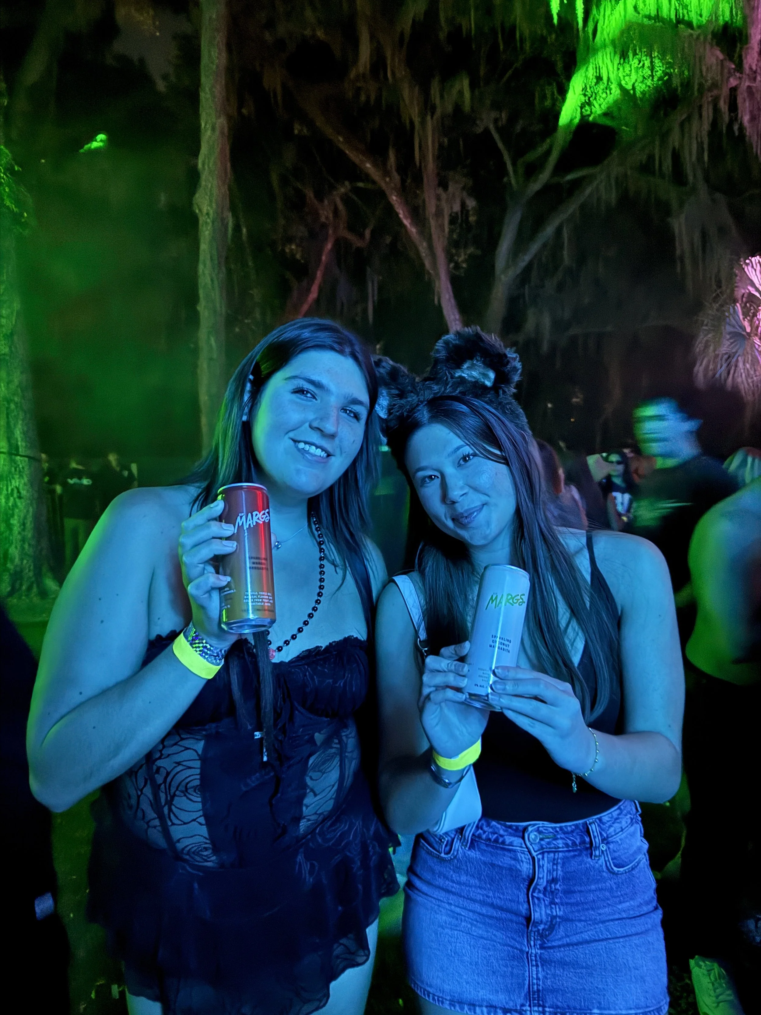 Two women holding cans and glasses, smiling at a party with colorful lighting and a jungle-themed background. One woman has bear ears headband and the other has dark hair and is wearing a black tank top.