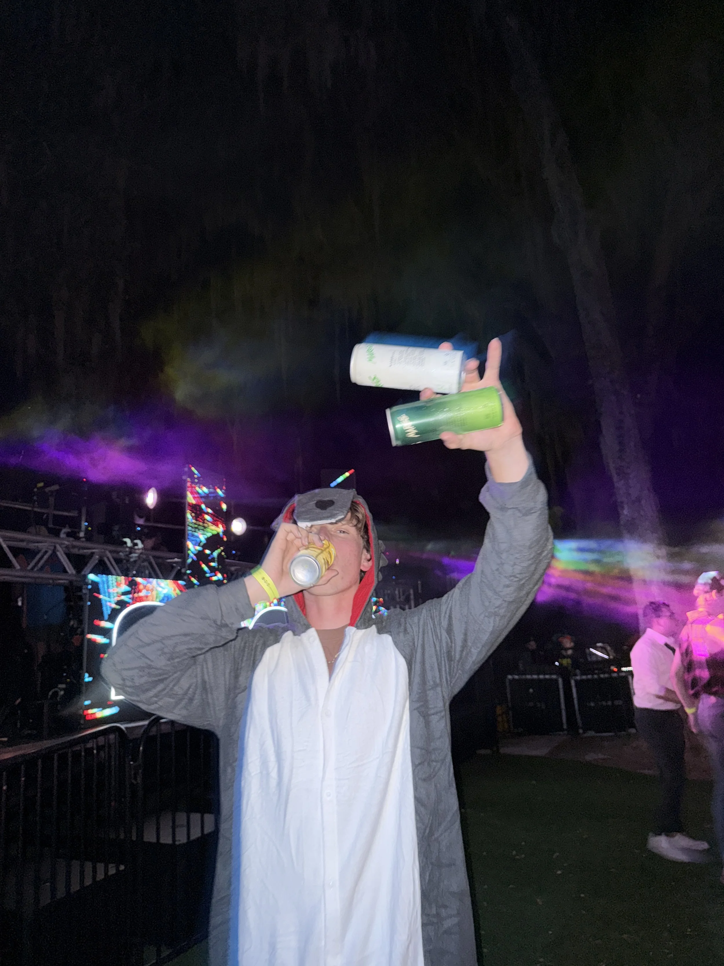 A young man wearing a hoodie and a ghost costume with googly eyes, drinking from a can and holding two other cans at a nighttime outdoor music event with colorful stage lights and other people in the background.