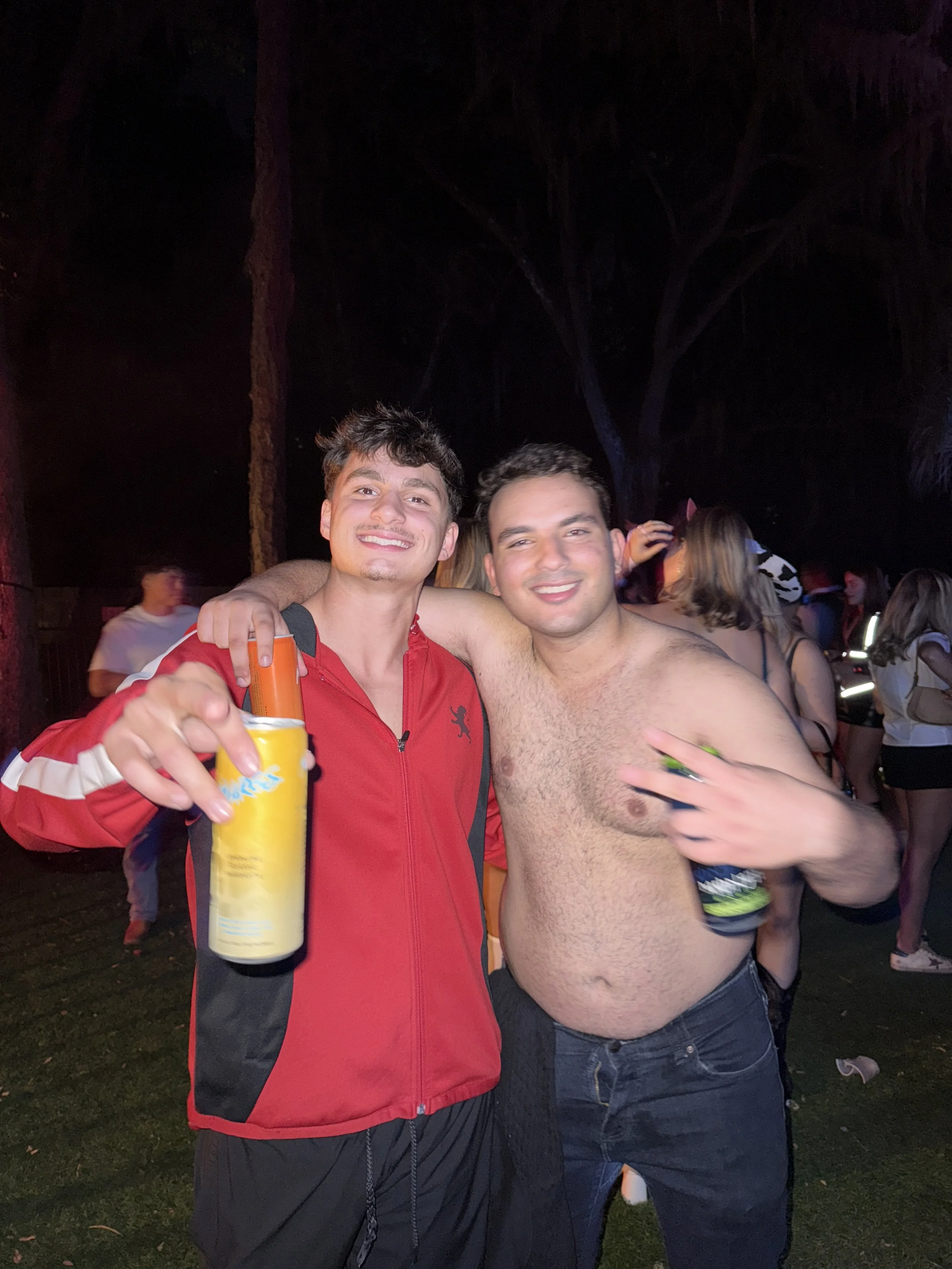 Two young men smiling, one wearing a red jacket and holding a drink, the other shirtless holding a can, at a nighttime outdoor event with a crowd in the background.