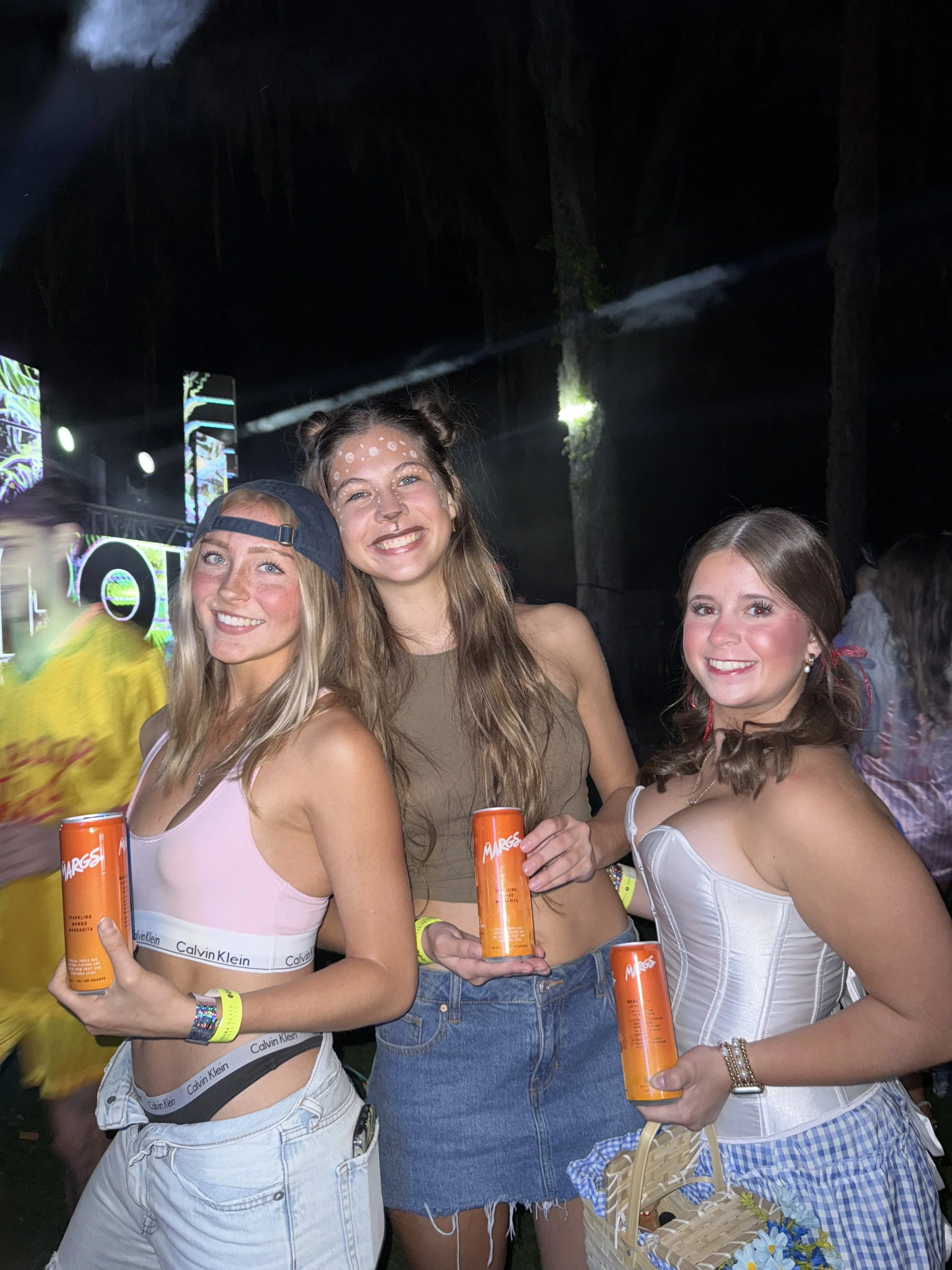 Three young women at a concert or festival, smiling and holding orange cans, with a stage and colorful lights in the background.