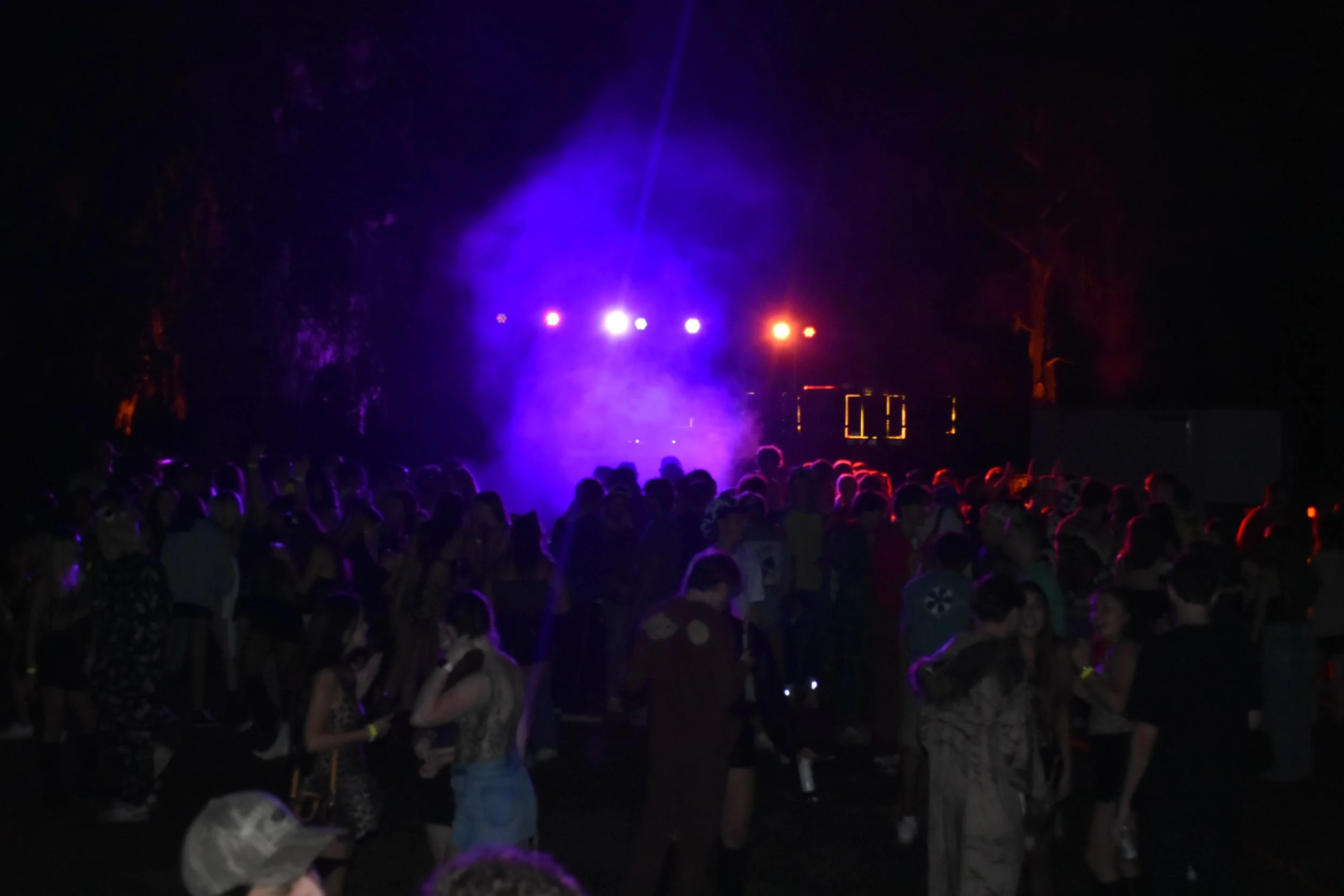 A crowd of people at a nighttime outdoor party with colorful stage lights and fog.