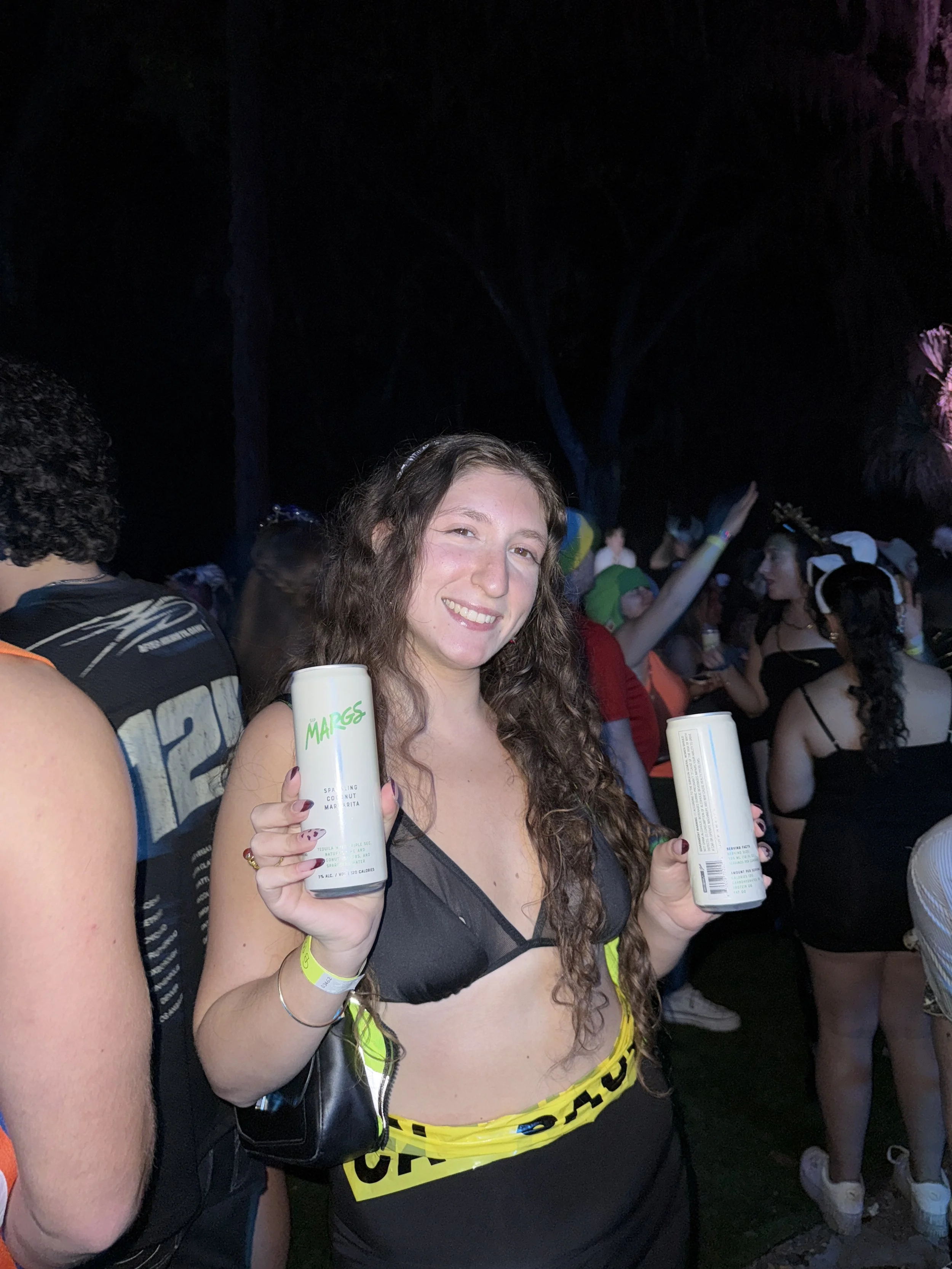 Young woman with long curly hair smiling at a nighttime outdoor party, holding two cans of drinks, wearing black and yellow clothing, surrounded by other partygoers.