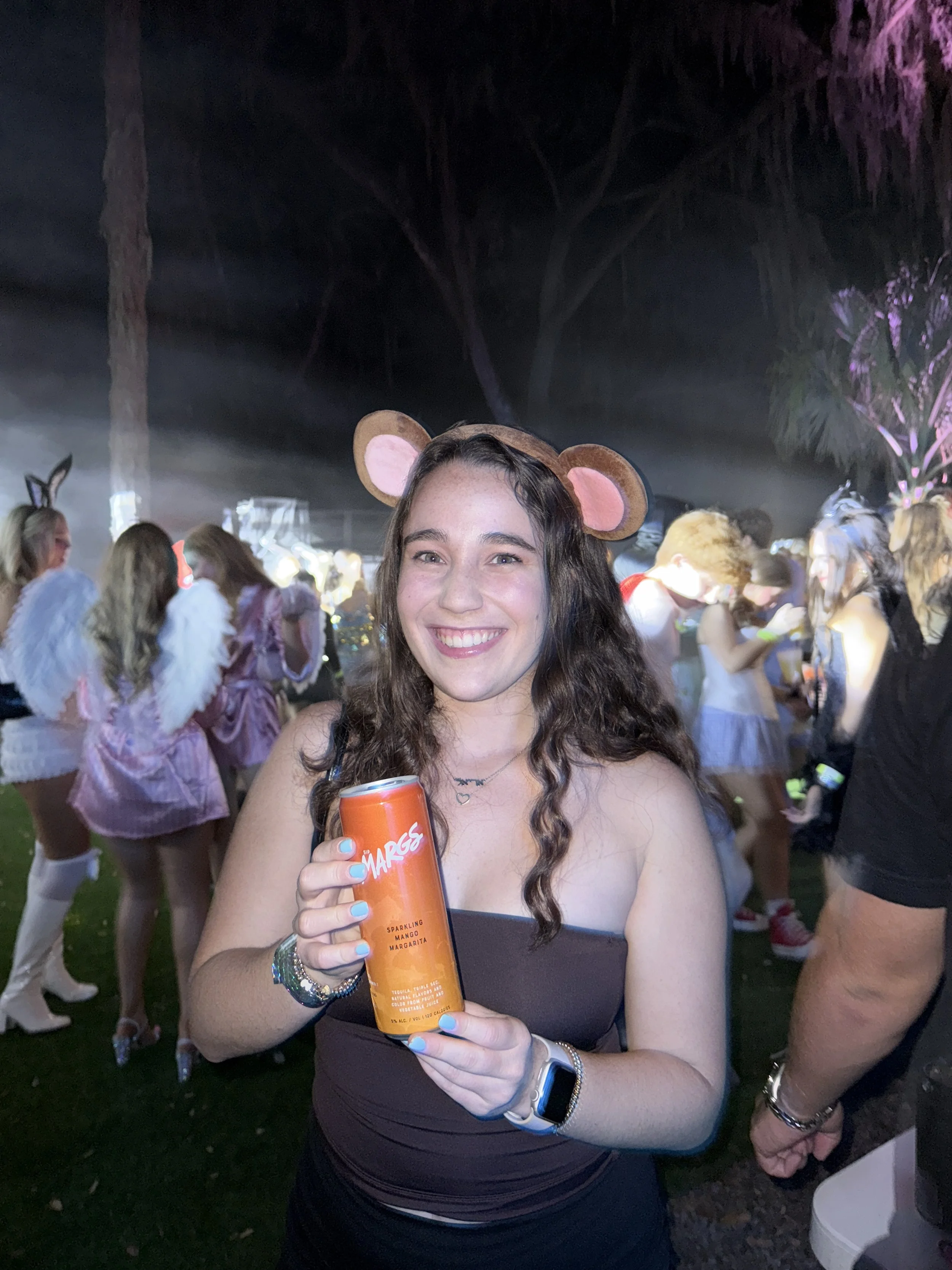 A young woman smiling at a party, holding a can of sparkling mango margarita. She is wearing a headband with mouse ears, a black strapless top, and has curly brown hair. The background is filled with people dressed in costumes, some with angel wings,