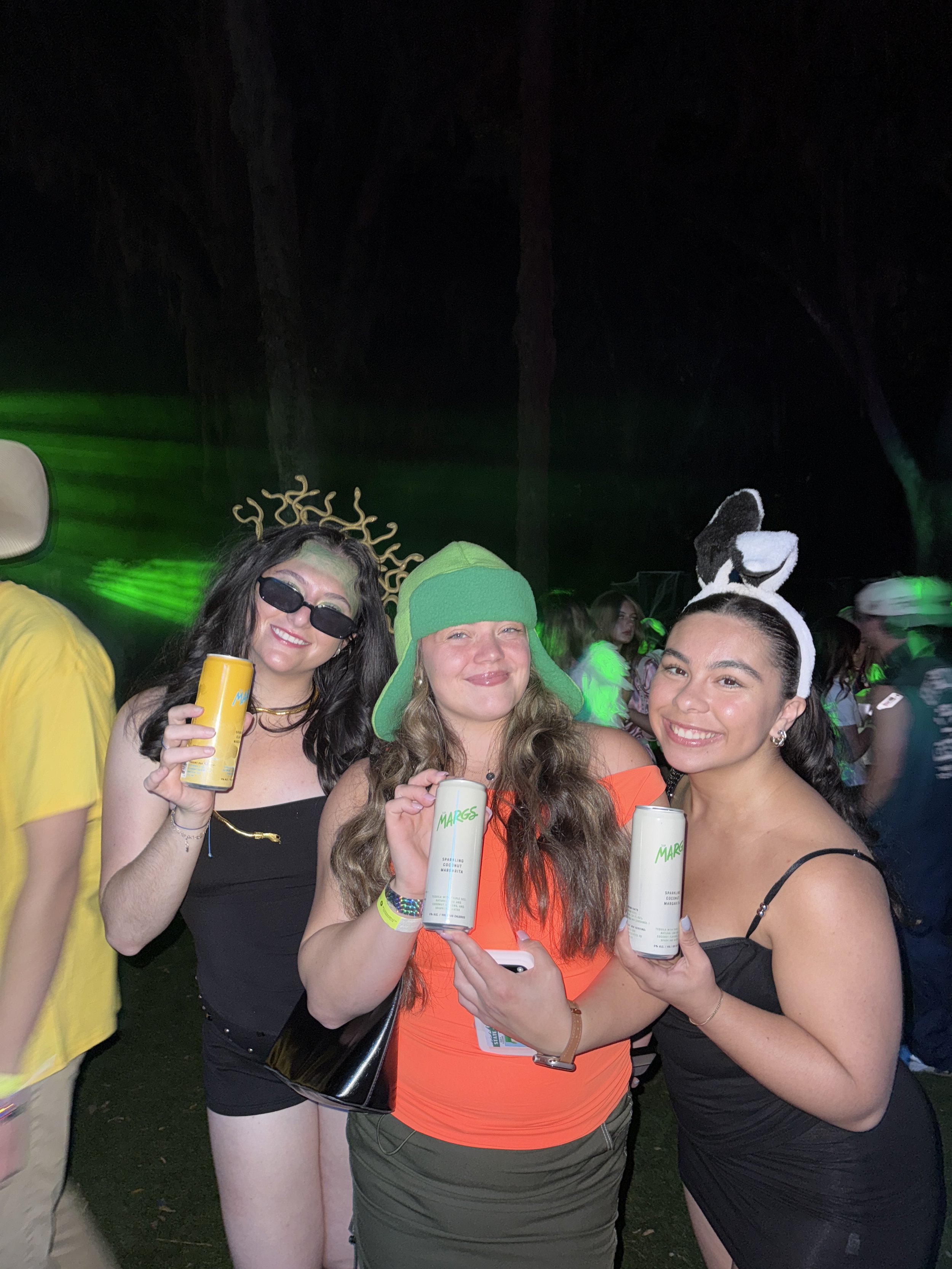 Three young women in costumes at an outdoor event, holding cans of sparkling water, smiling, with green lighting and other people in the background.
