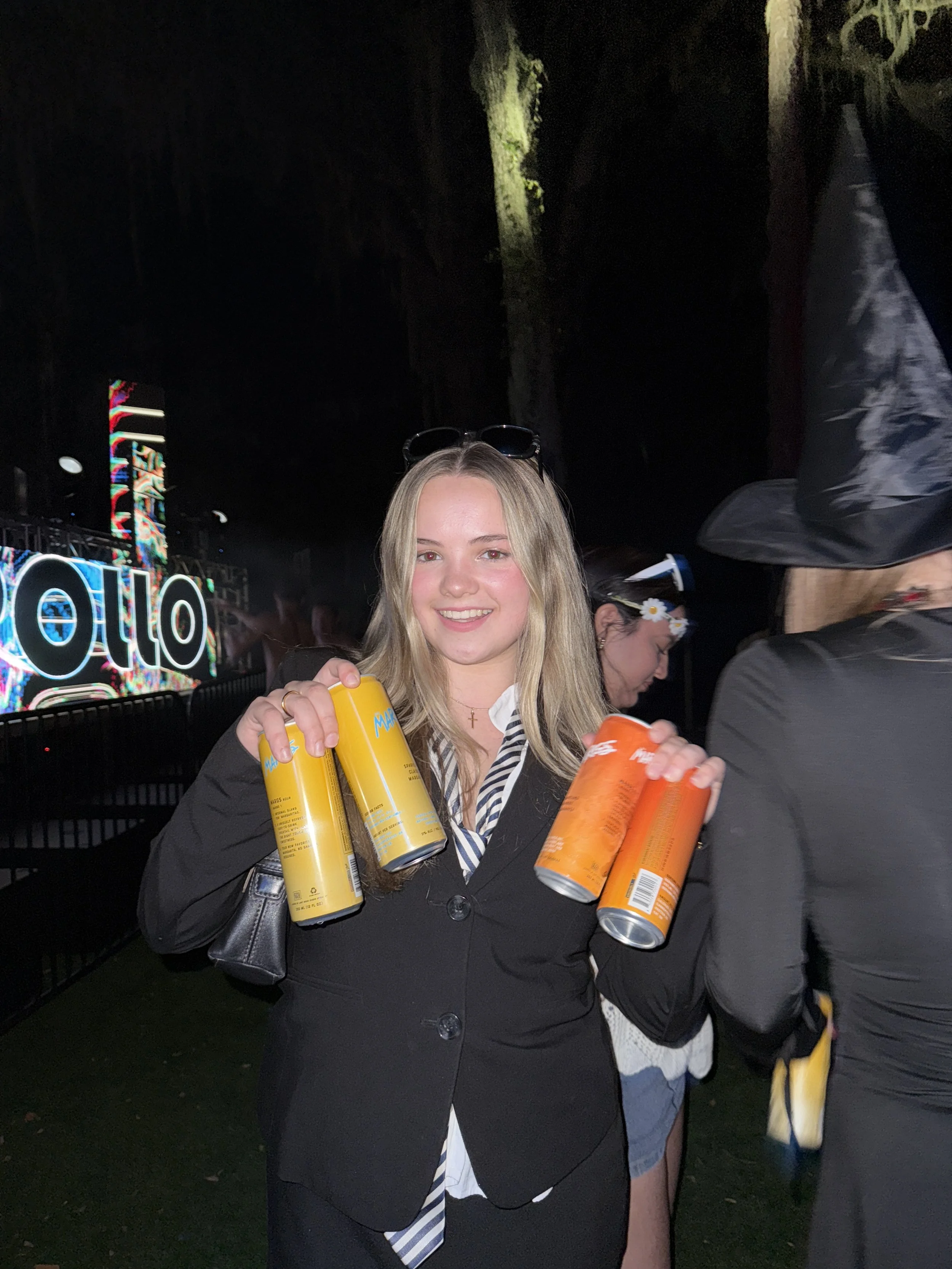 A smiling young woman with blonde hair holding four cans of Mana energy drink at a lively event with a stage and colorful lights in the background.
