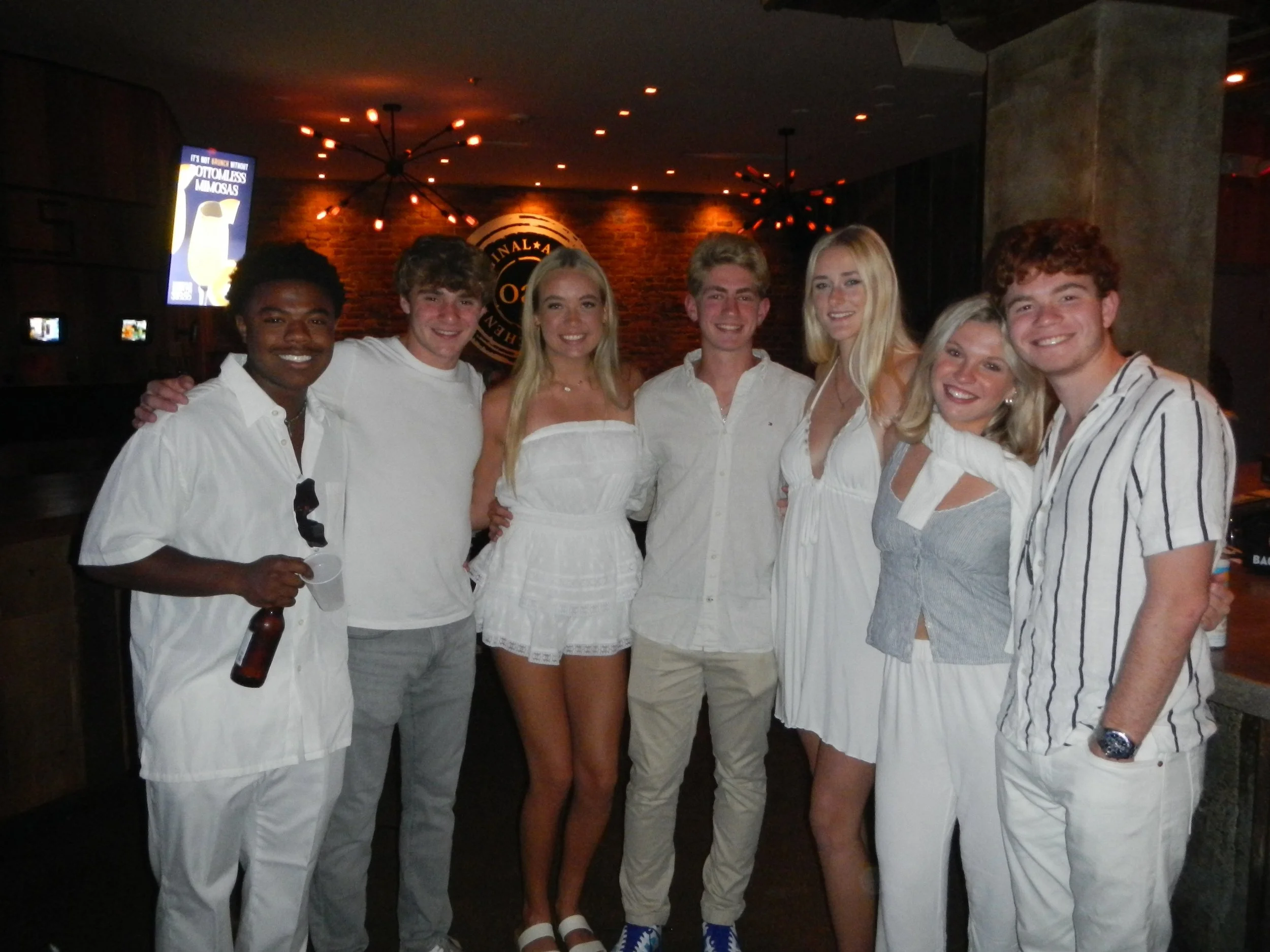 Group of eight young adults posing together in a bar or restaurant, dressed in white or light-colored clothing, smiling and standing close together.