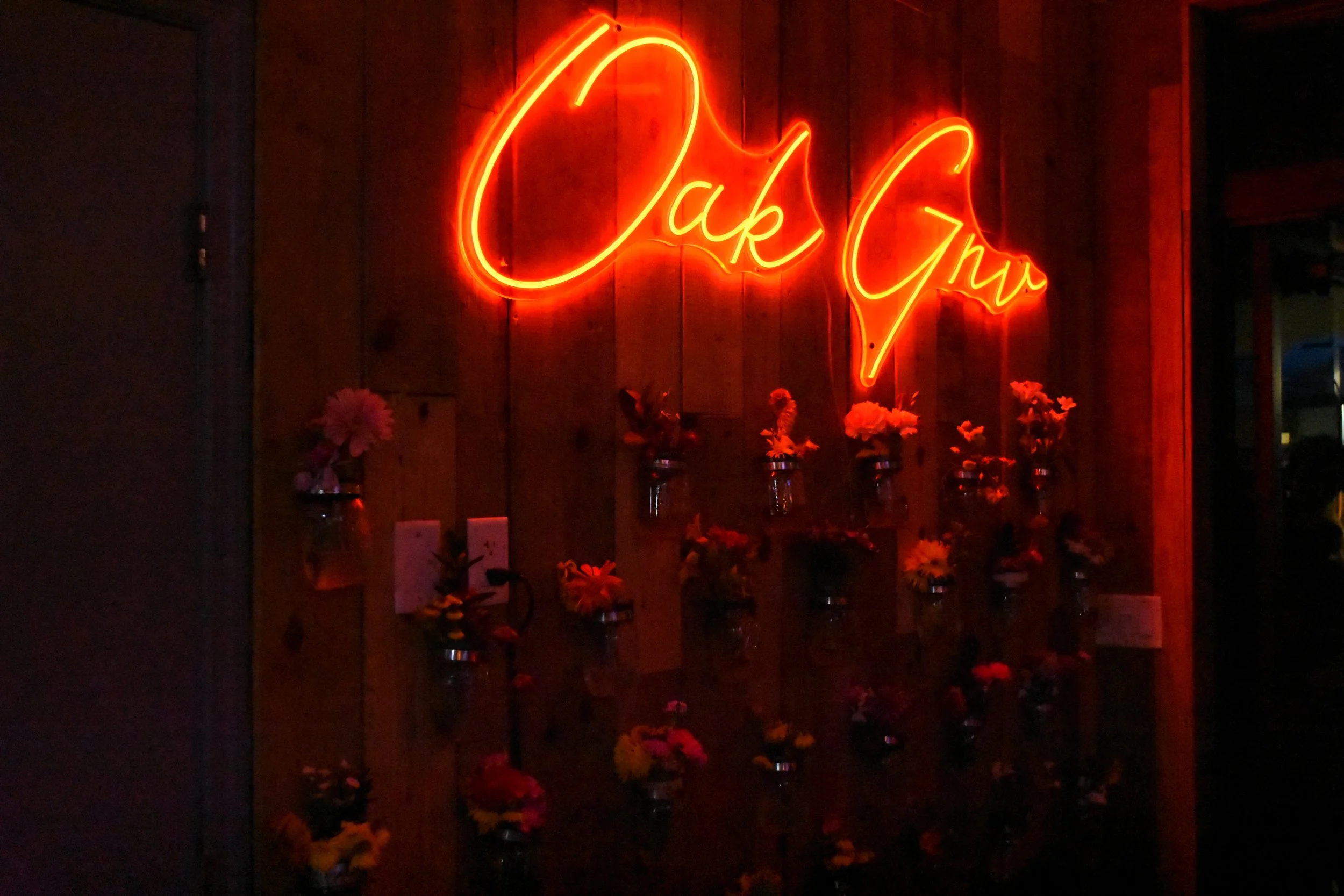 Neon sign reading 'Oak GNV' on a wooden wall, decorated with small flower vases.