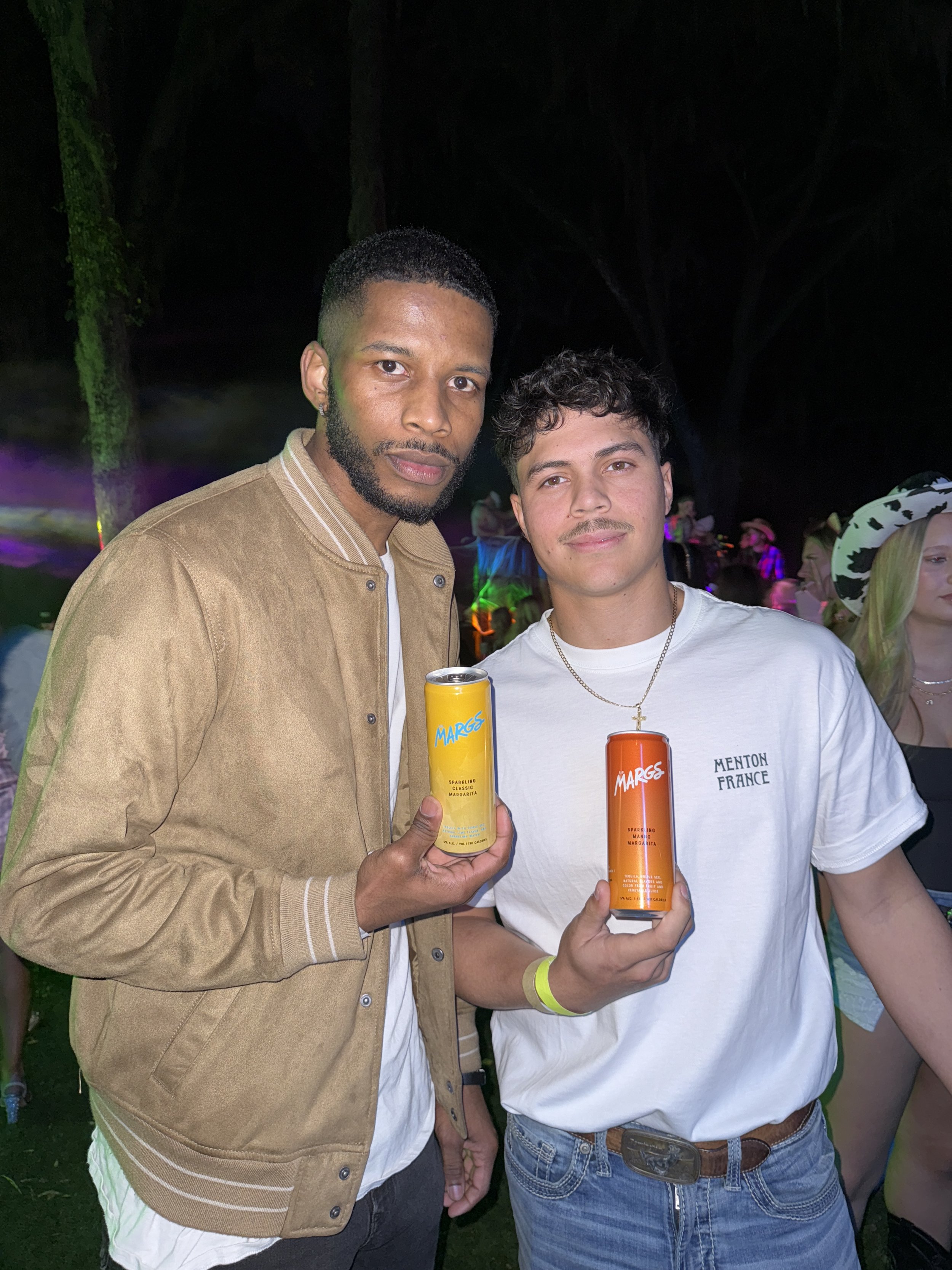Two young men holding cans of Marges beverages at an outdoor event at night, with colorful lights and other people in the background.