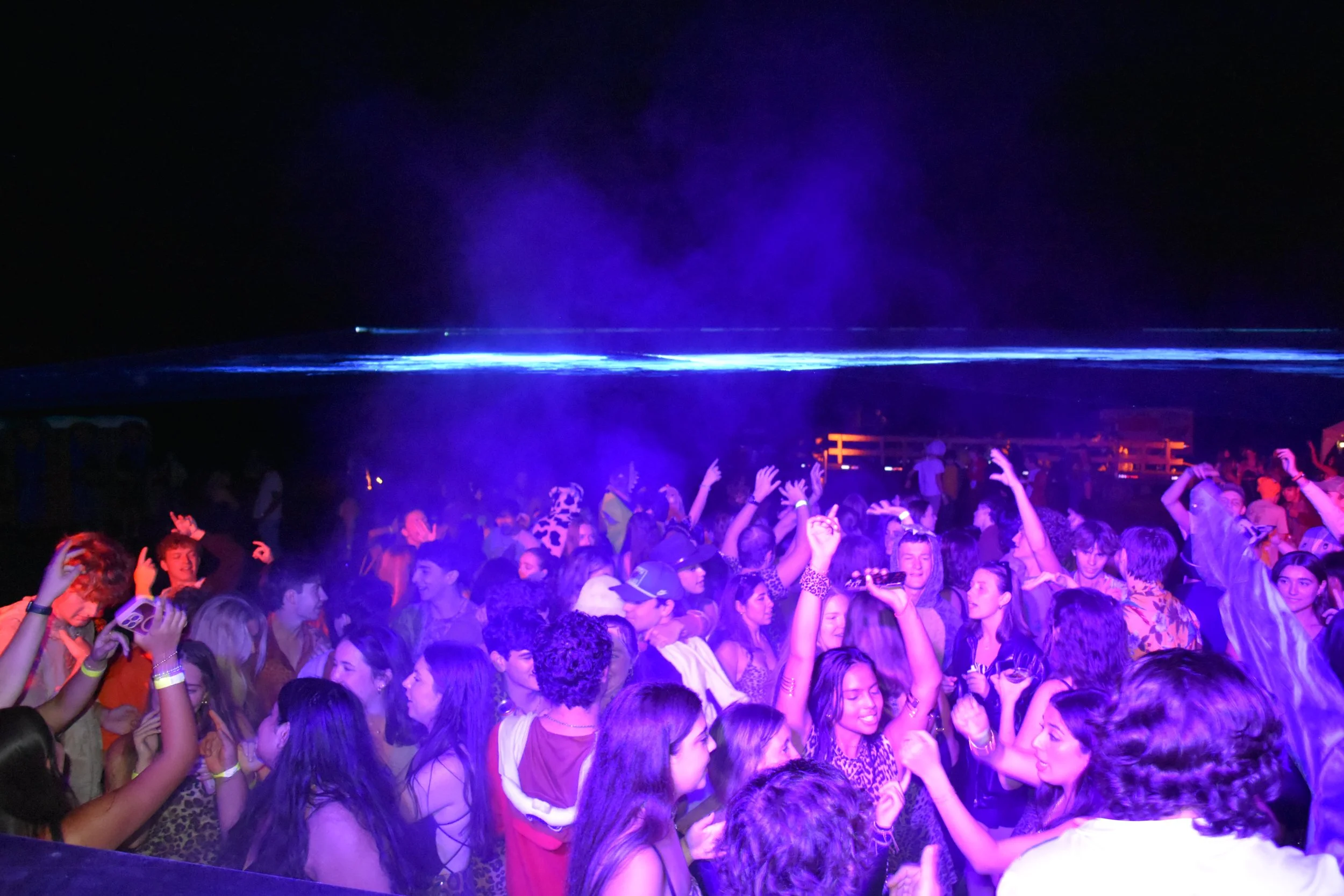 A large group of people dancing and enjoying music at a party or concert with purple and blue lighting effects.