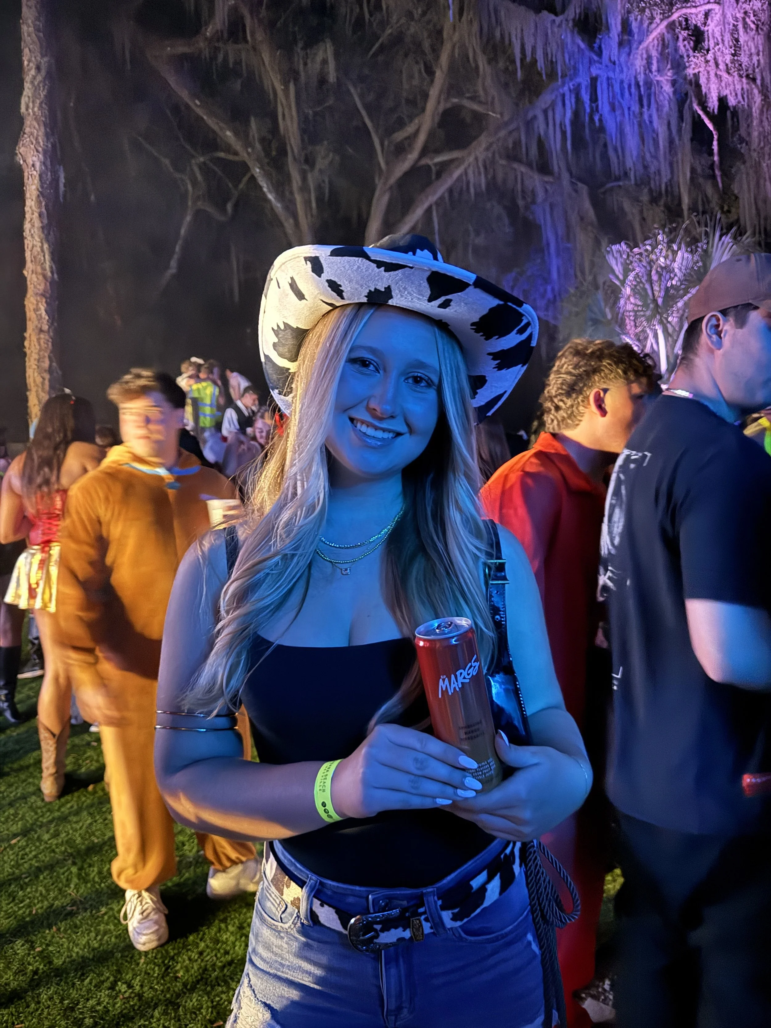 A young woman wearing a cow print hat and black top, holding a can of Marges soda, at a nighttime outdoor event with colorful lighting and a crowd of people in the background.