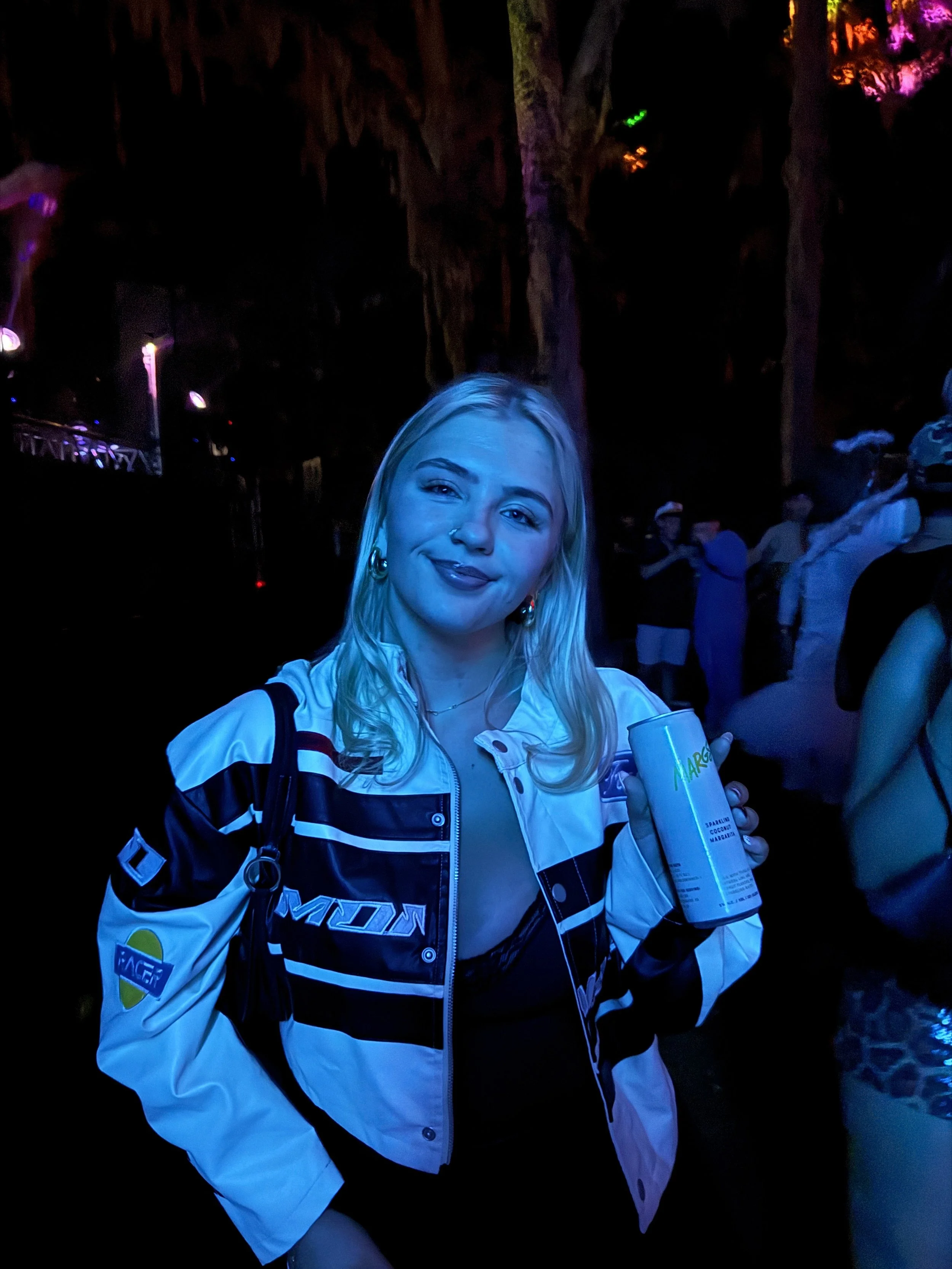 A young woman with blonde hair, wearing a white and black jacket, smiling while holding a can at a nighttime event with colorful lighting and other people in the background.