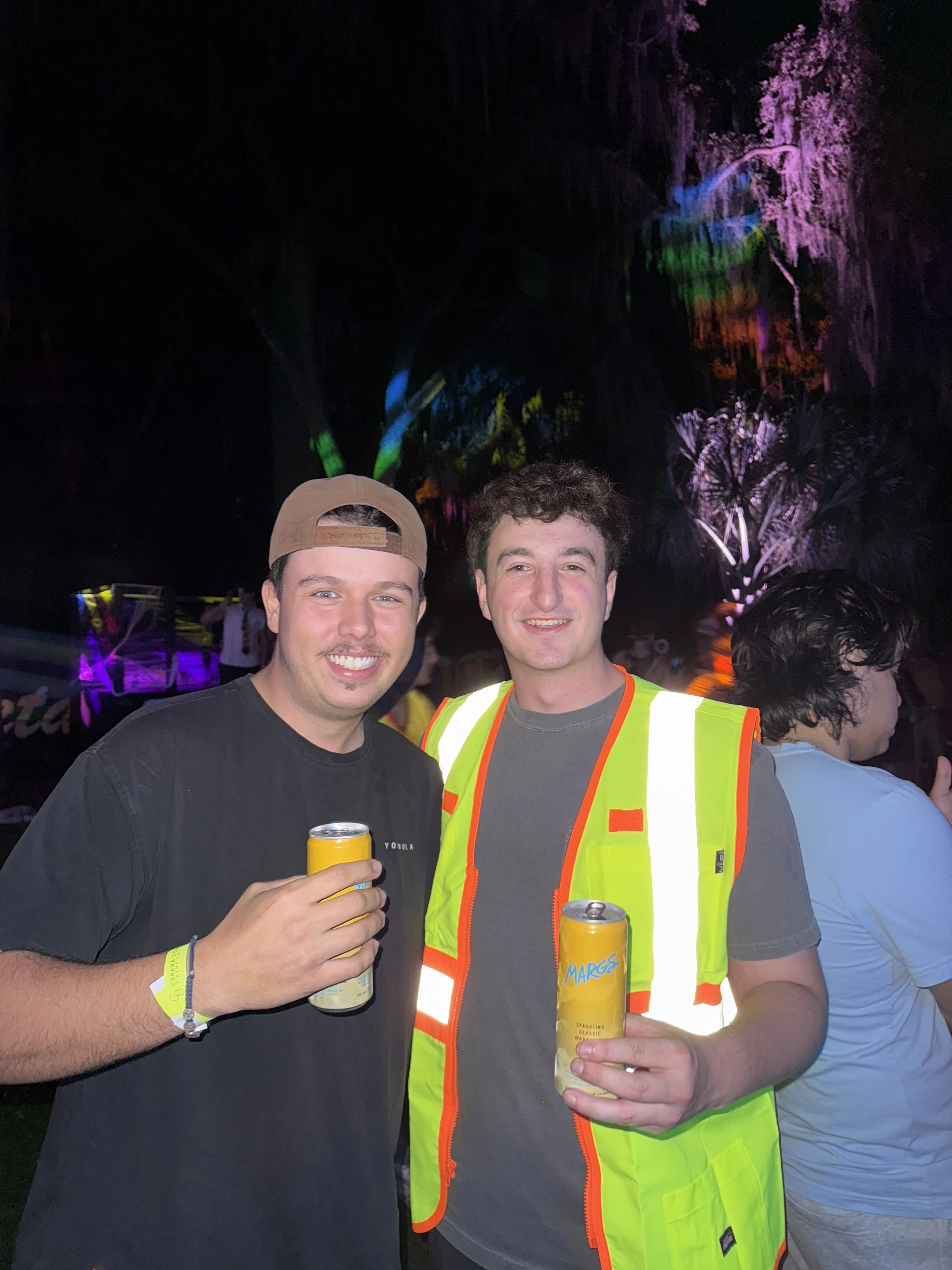 Two young men smiling at night, holding cans of Marg’s sparkling wine, with colorful lights and trees in the background, one wearing a high-visibility vest and the other wearing a backward cap.