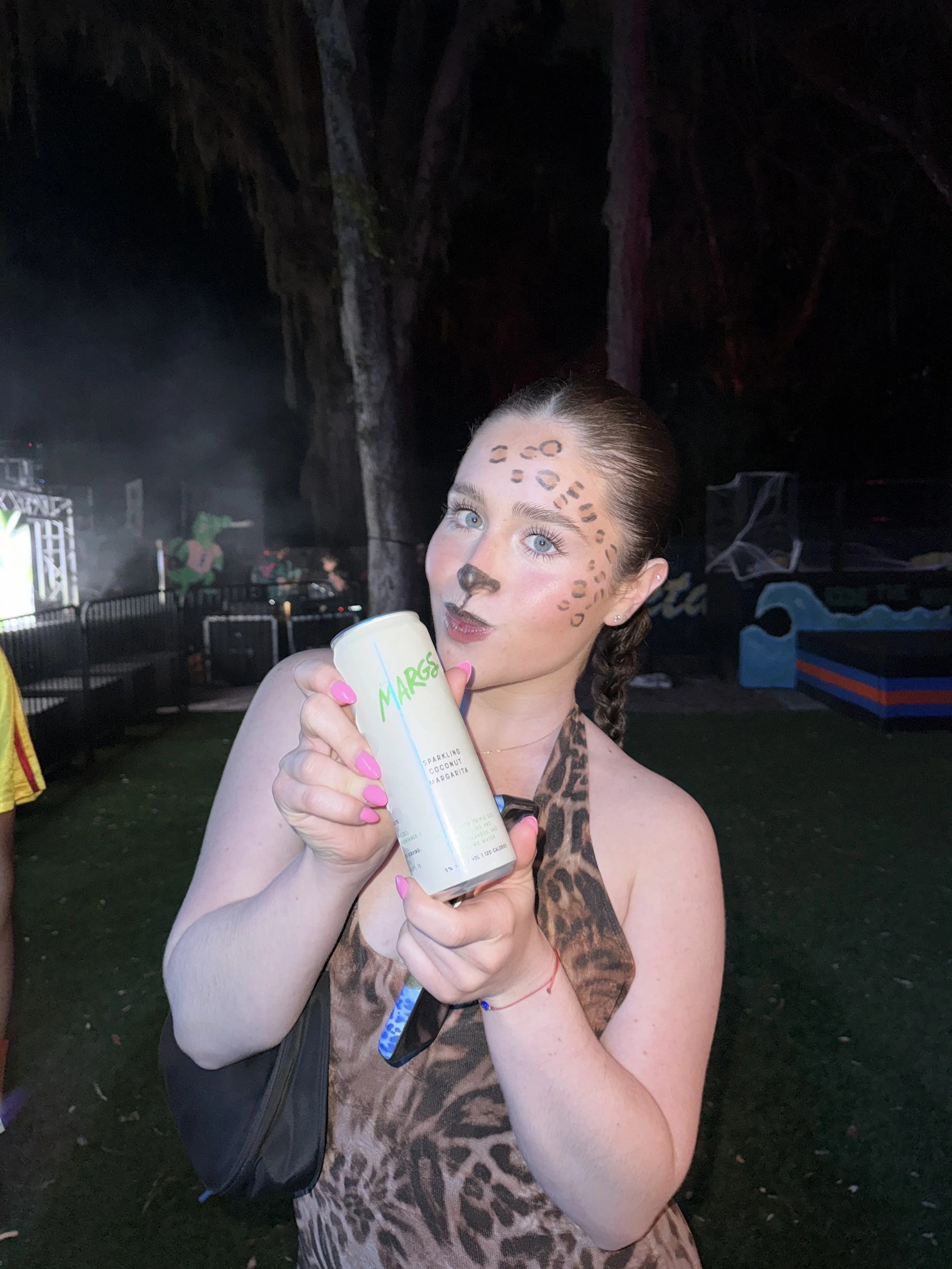 A woman with face makeup resembling a cheetah holding a can of sparkling coconut margarita at an outdoor event at night.