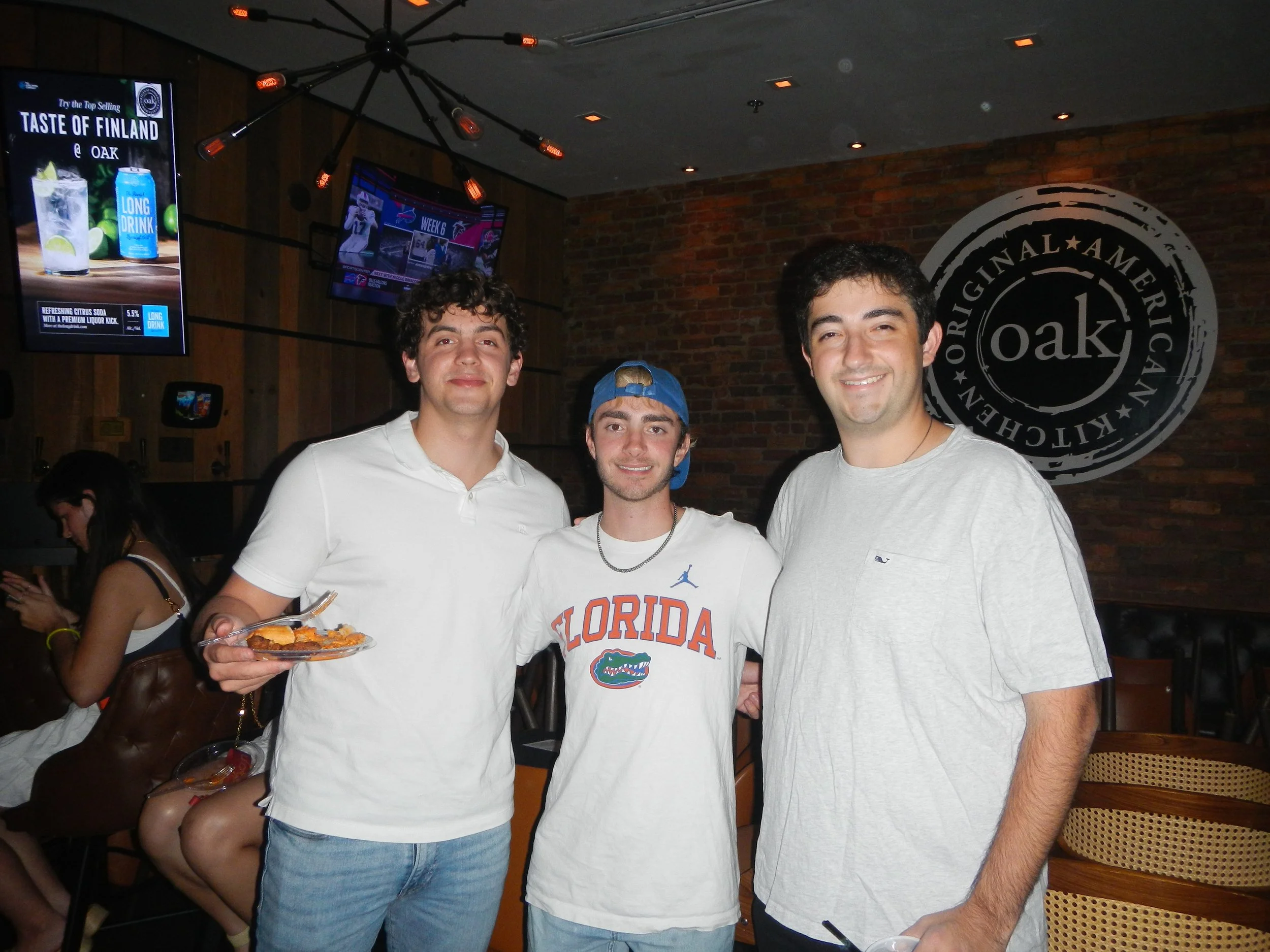 Three young men smiling at a social gathering in a restaurant, one holding a plate of food. They are standing in front of a brick wall with a large circular logo that says 'oak' and 'original American Kitchen.'