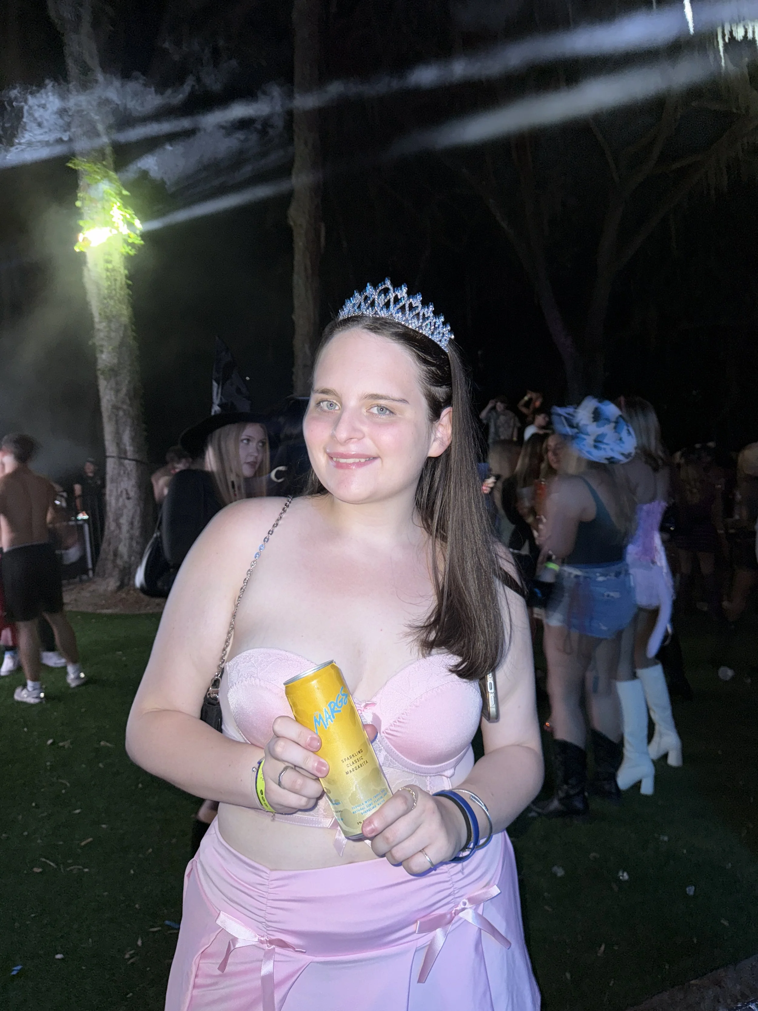 A young woman with long brown hair, wearing a silver tiara, a pink strapless dress, and multiple bracelets, stands outdoors at night holding a canned beverage at a lively party.