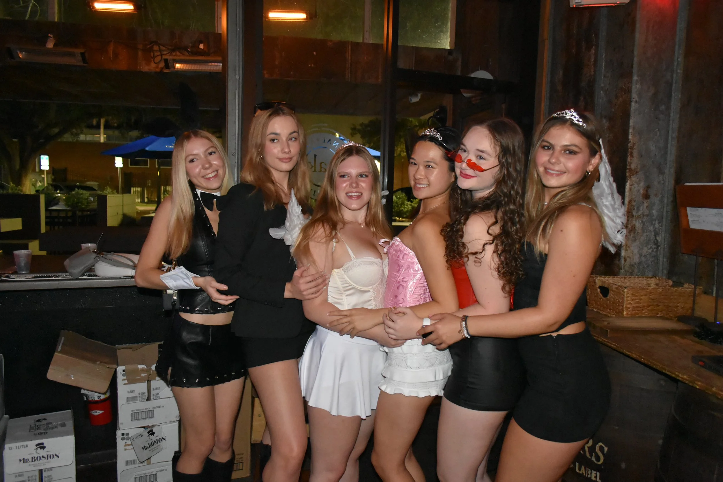Six women dressed in party costumes, wearing tiaras and accessories, pose together at a social gathering in a wooden decorated venue.