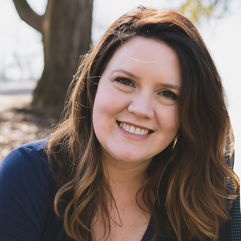 Close-up of a young woman with long brown hair smiling outdoors near a tree.