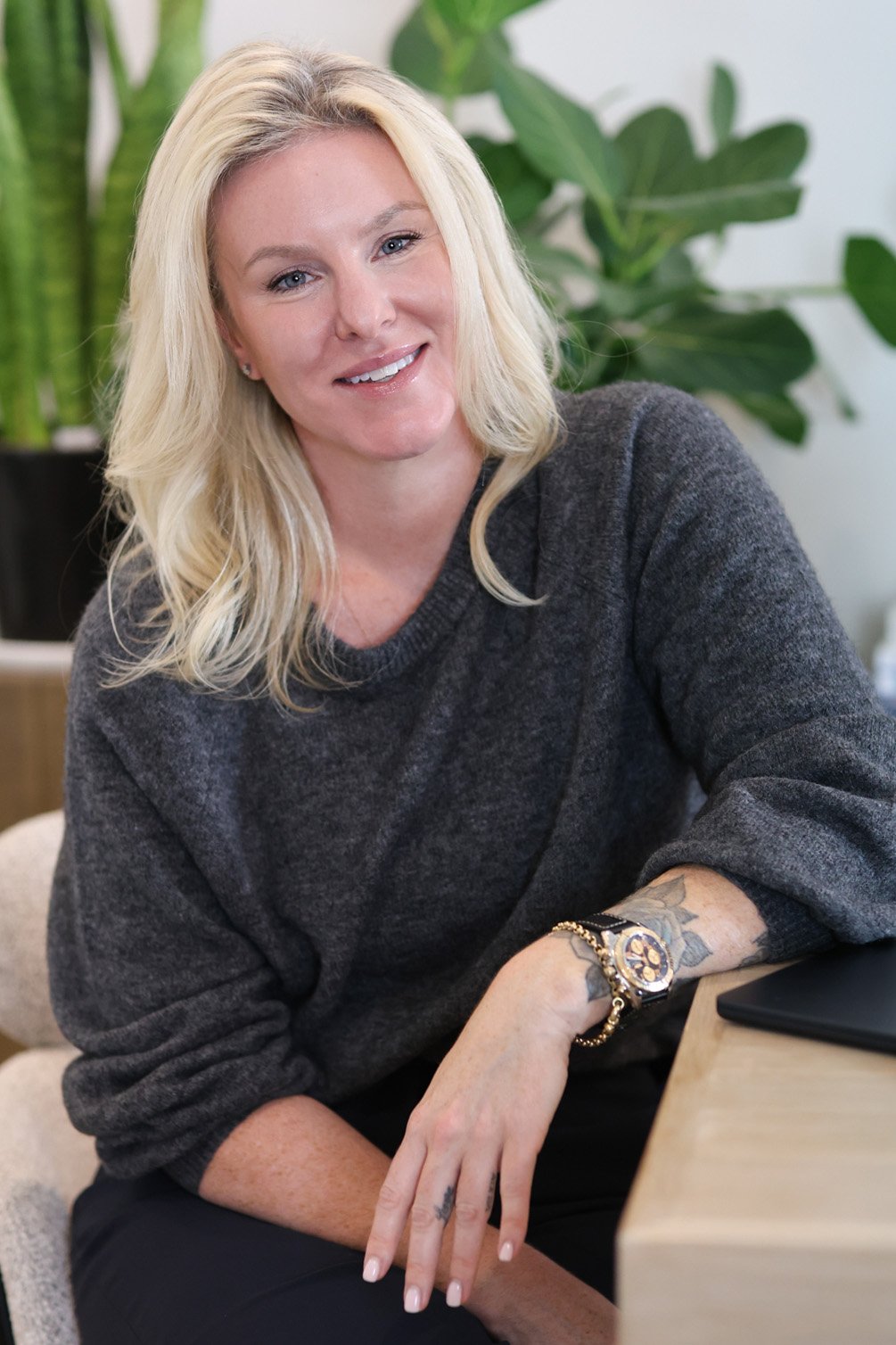 A smiling woman with long blonde hair in a dark gray sweater, sitting at a table with plants in the background.