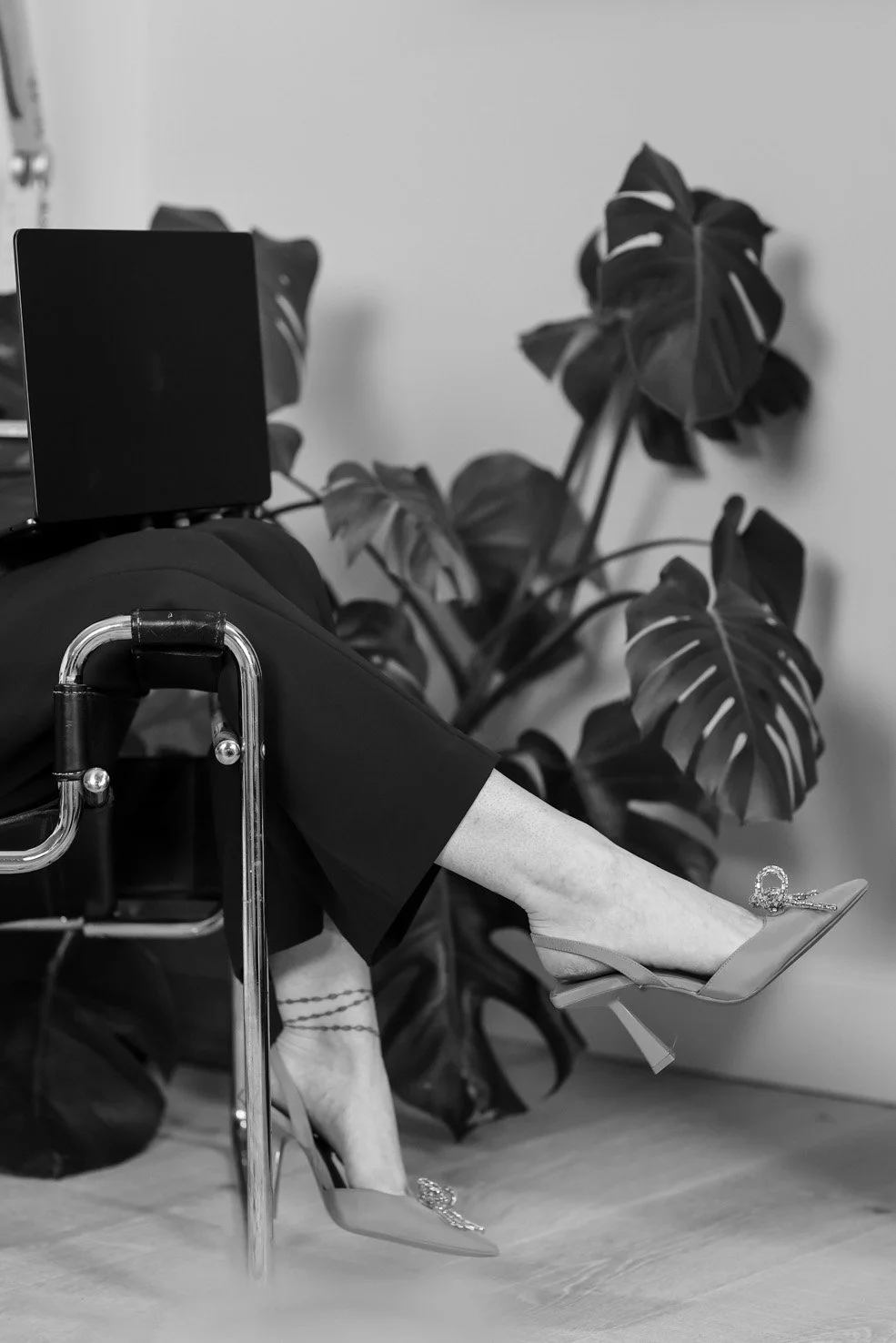 A person in a wheelchair with legs crossed at the ankle, wearing high heels with decorative stones, and black pants, positioned in front of large houseplants with monstera leaves.