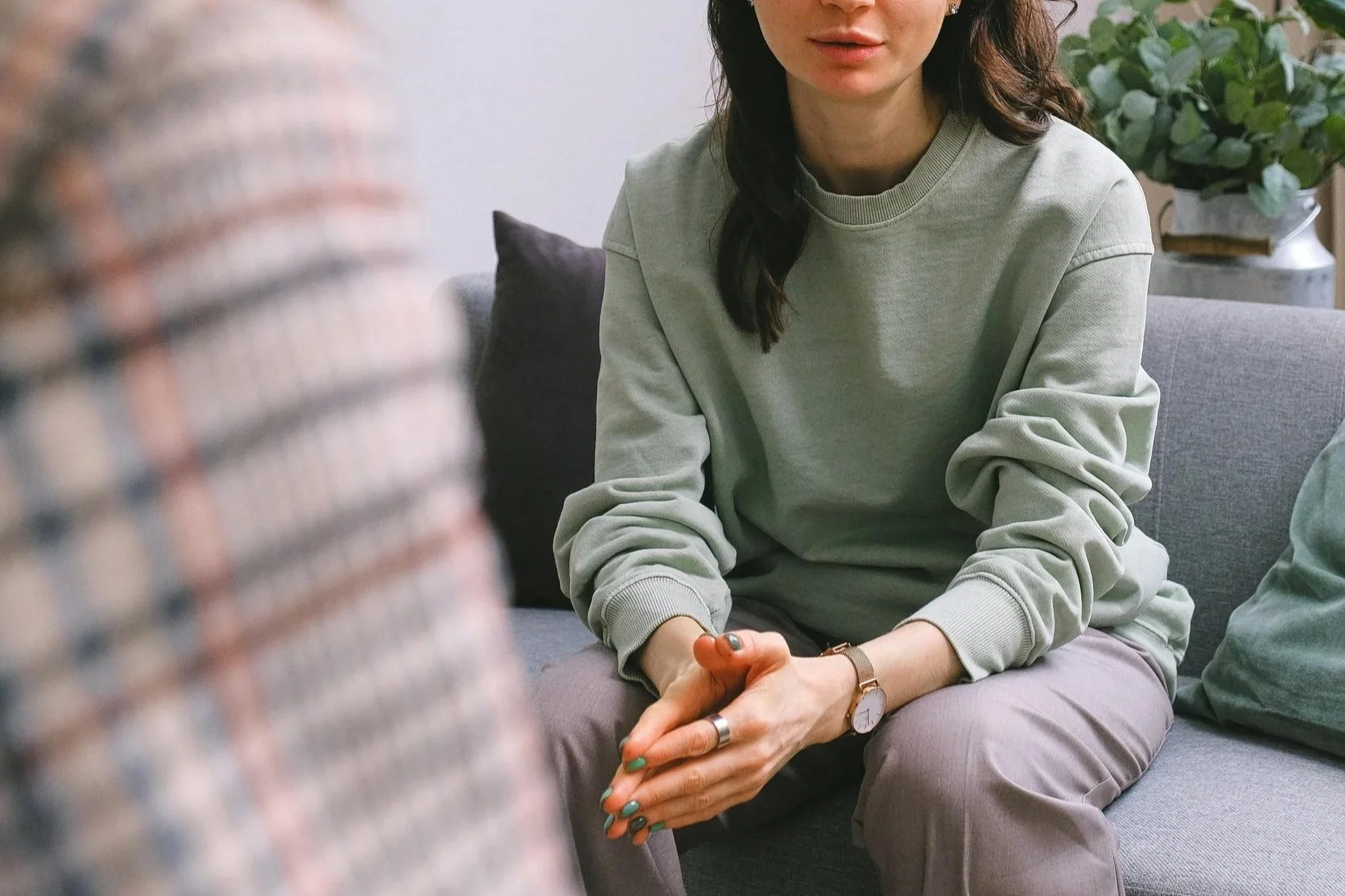 A woman sitting on a sofa with her hands clasped together, wearing a pale green sweatshirt, gray pants, and a watch, with a potted plant in the background.