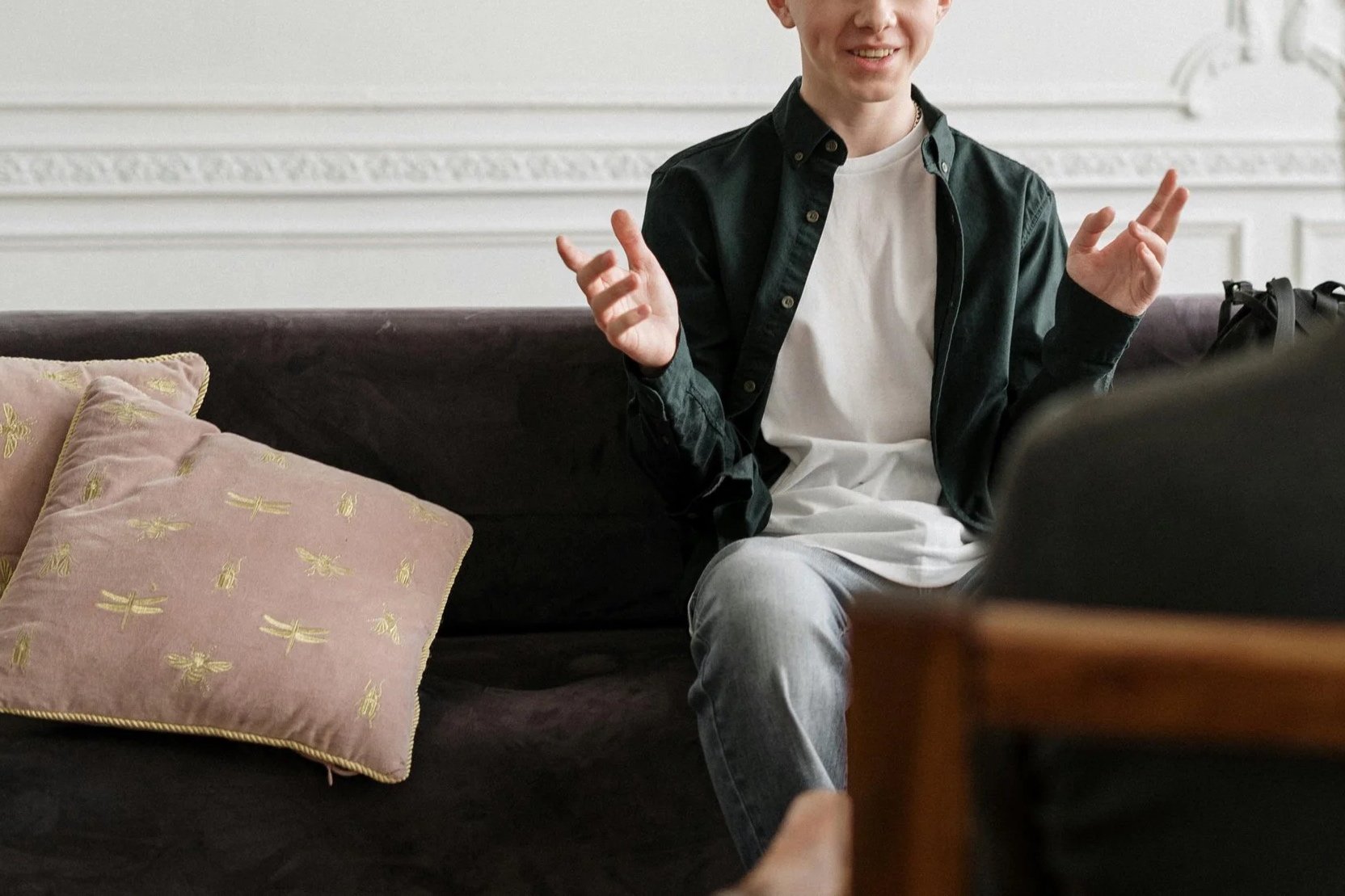 A young man sitting on a dark sofa with pink pillows, wearing a black shirt over a white t-shirt, smiling and gesturing with his hands.