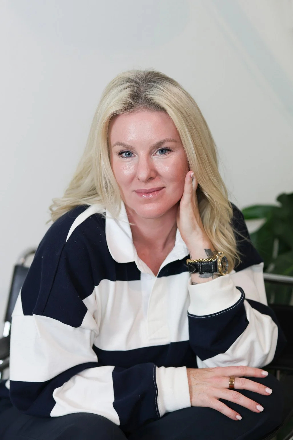 A blonde woman with blue eyes, wearing a striped black and white shirt, sitting indoors against a white wall. She has a watch and ring on her left hand, with her hand touching her face, and is smiling slightly at the camera.