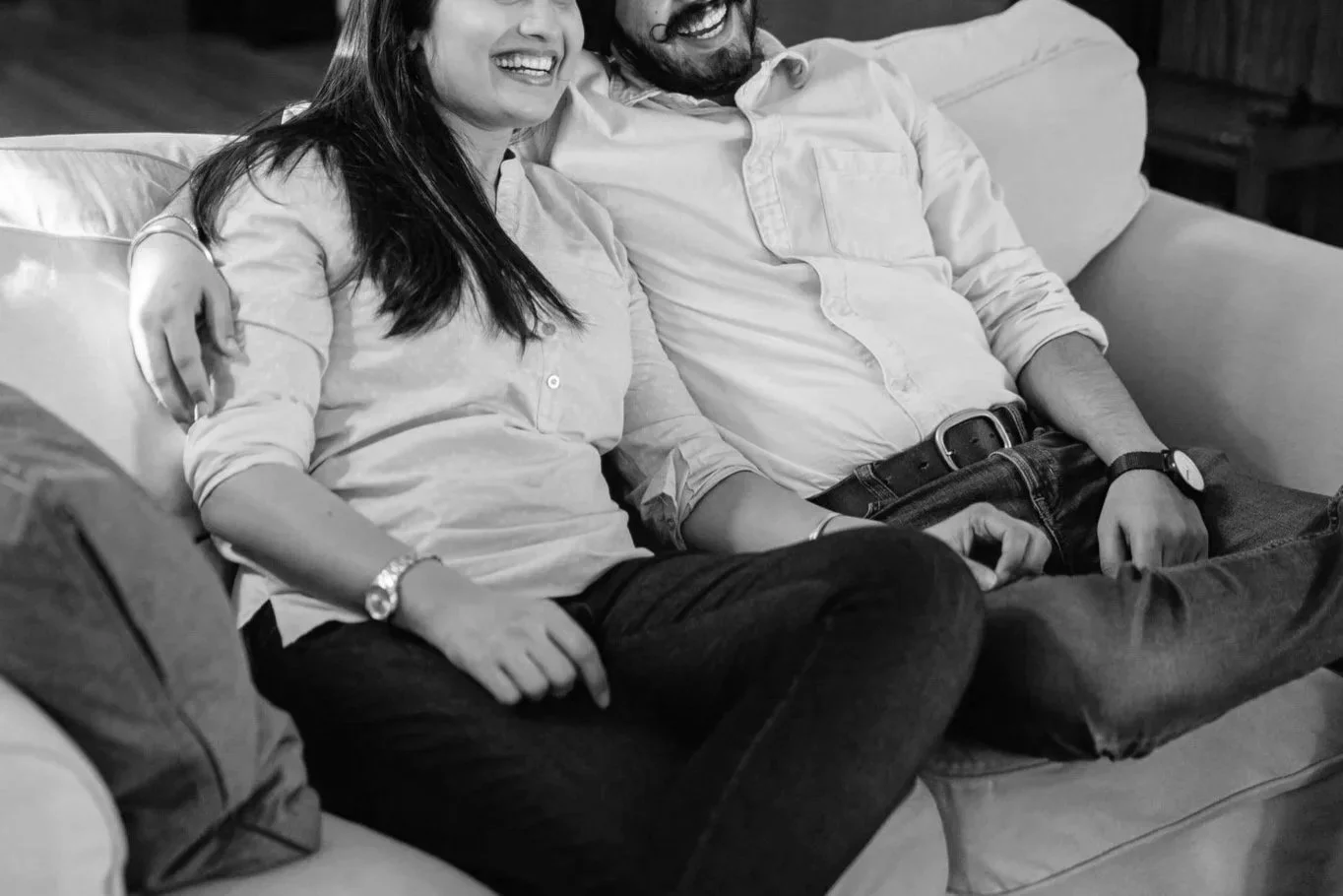 A black-and-white photo of a smiling woman and man sitting close together on a sofa, holding hands and showing affection.