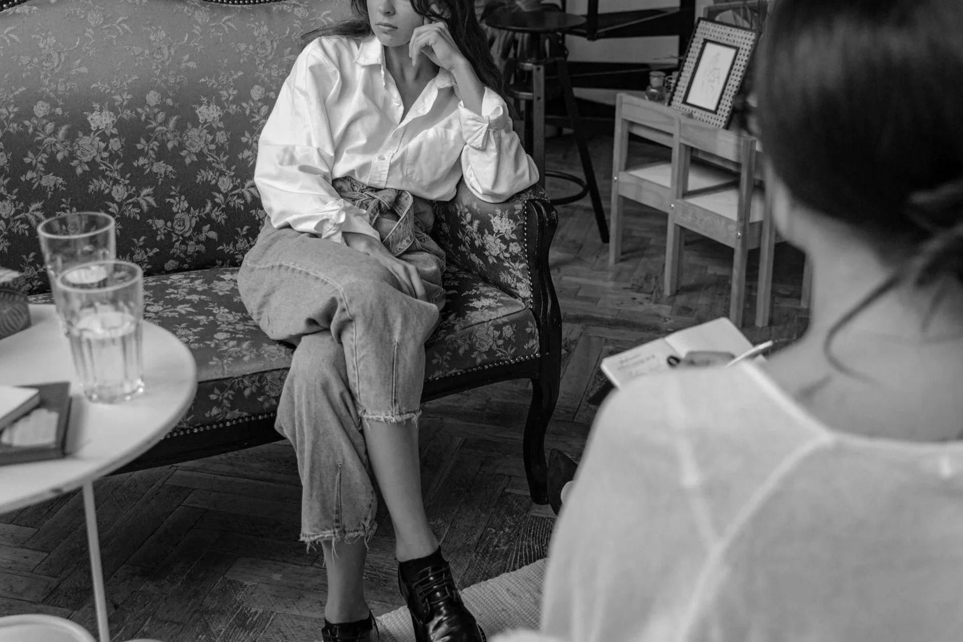 A woman sitting on a floral-patterned sofa appears to be in a counseling or therapy session. She wears a loose white shirt, high-waisted jeans, and black shoes, with her hand resting on her chin, looking contemplative. In front of her, another person, likely a therapist, is seen partially, holding a notepad and pen.