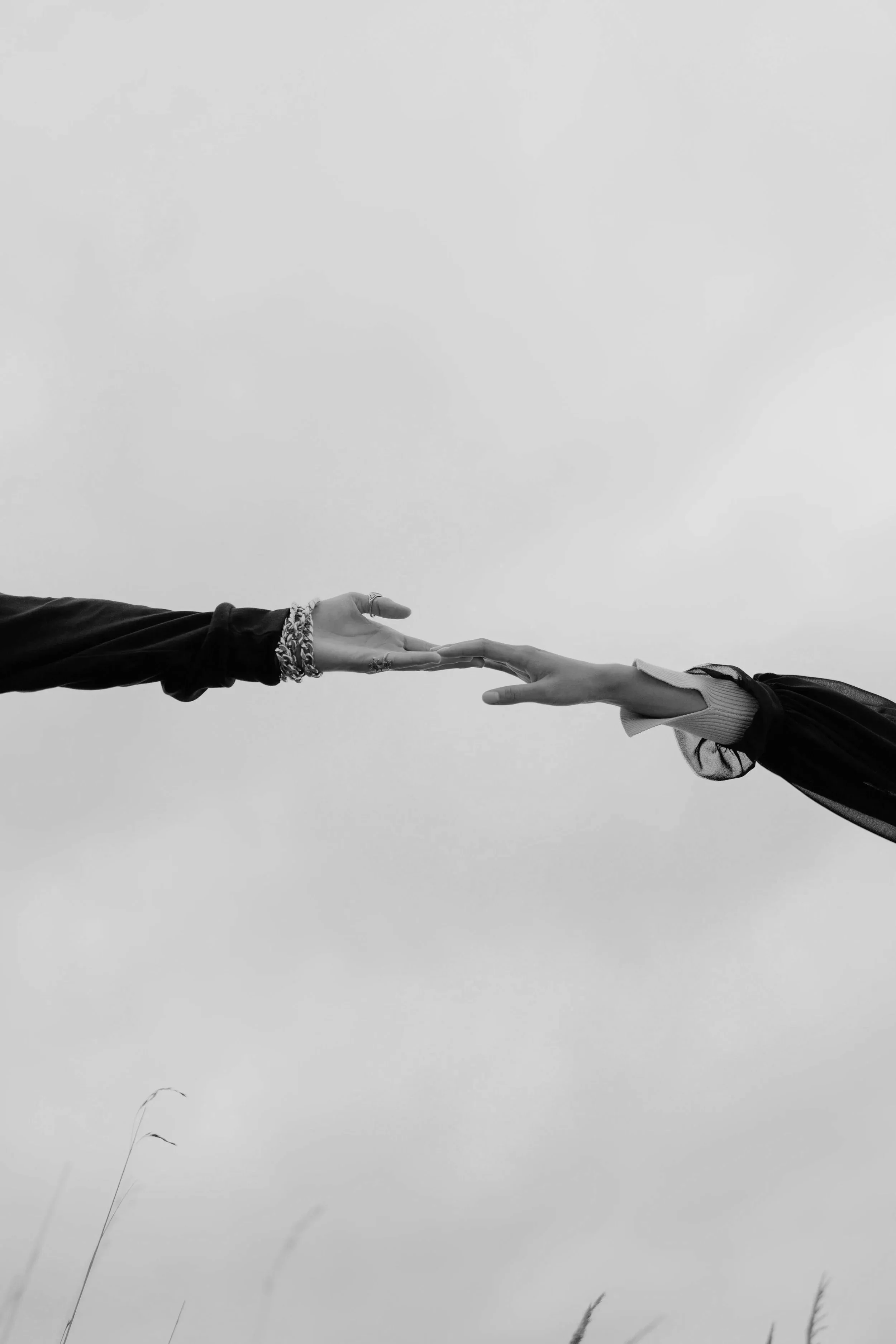 Two hands reaching towards each other, with fingers touching, against a cloudy sky background in black and white.