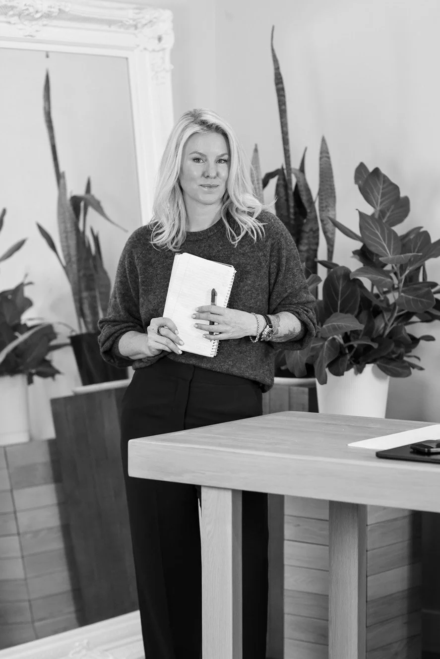 A woman with blonde hair holding a notebook and pen, standing next to a desk with plants in the background.
