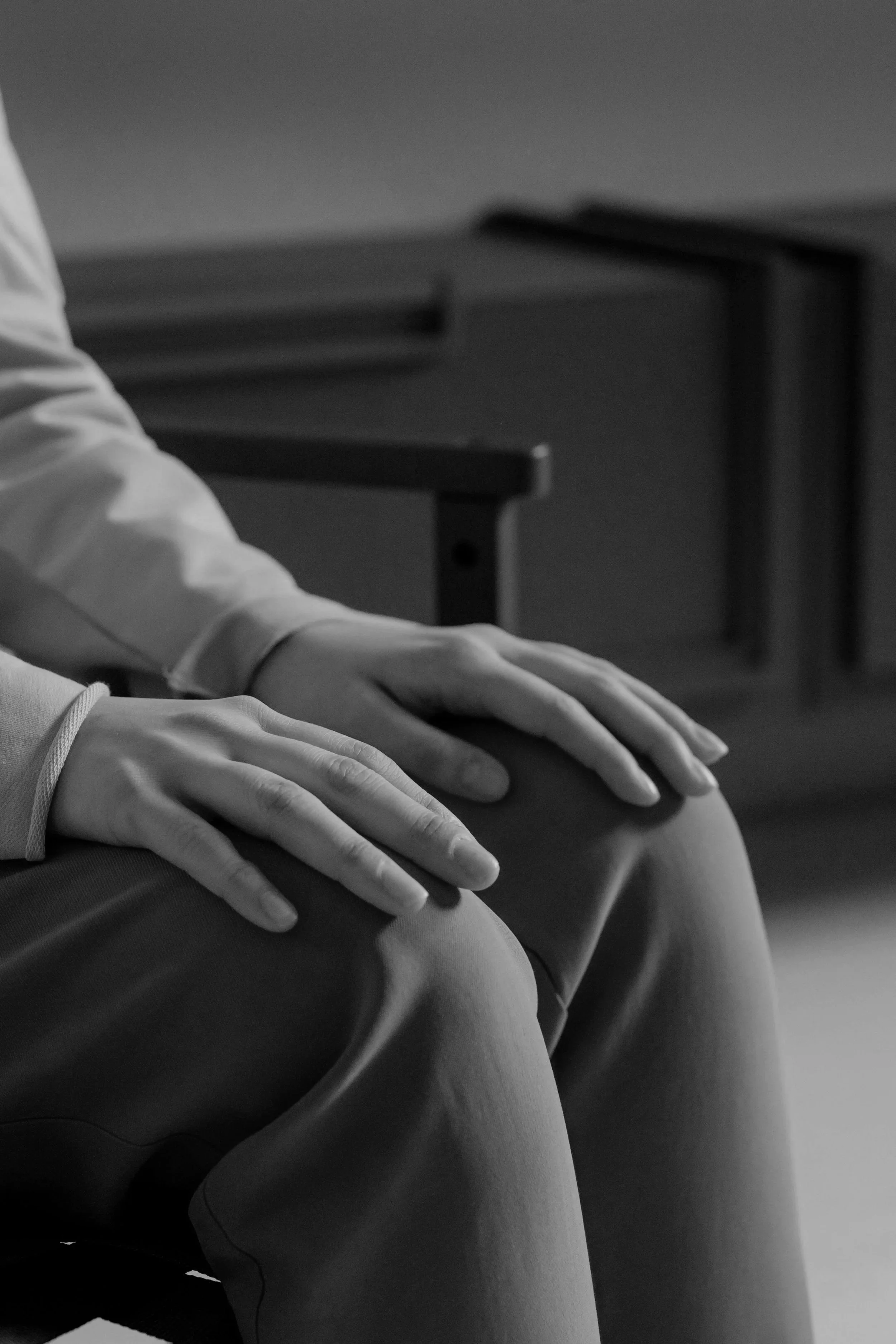 Black and white photo of a person's hands resting on their lap while sitting, with a table or desk in the background.