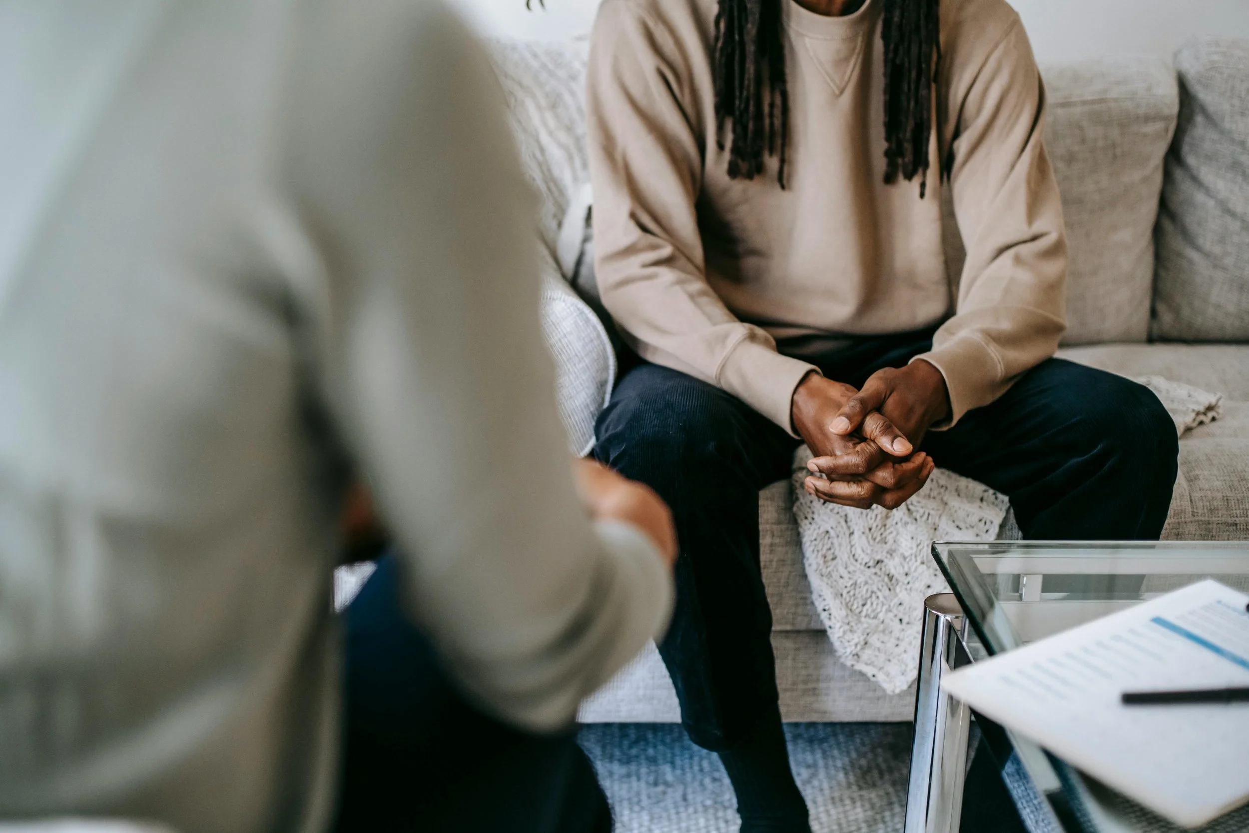 A person sitting on a beige sofa, clasping their hands, engaging in conversation with another individual in a beige sweater, during a counseling or therapy session in a living room.
