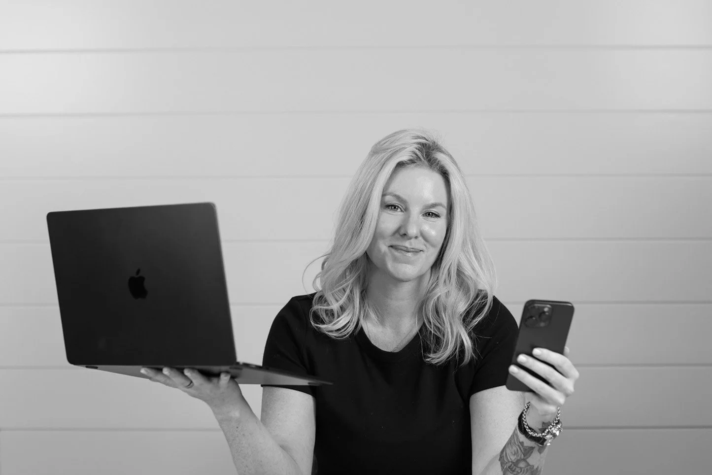 Black and white photo of a woman smiling, holding a smartphone in her right hand and a laptop in her left hand, standing against a plain wall.