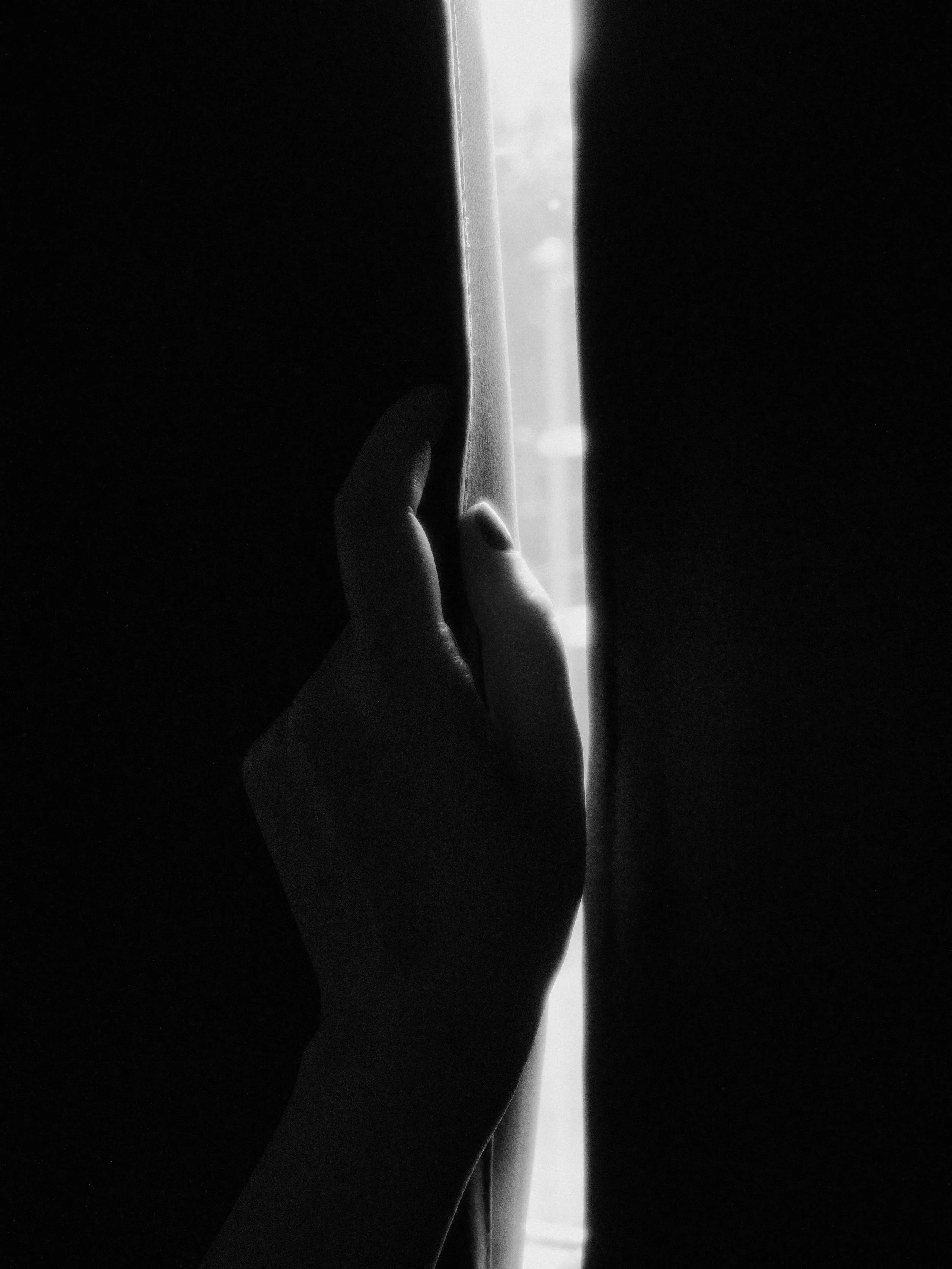A hand is pulling back a curtain to reveal a bright window, creating a contrast between darkness and light.
