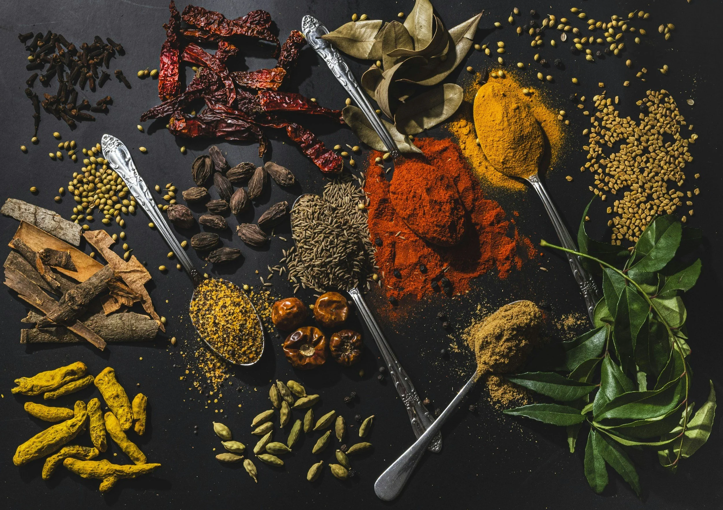 Assorted spices on a dark surface, including dried chili peppers, turmeric root, cumin seeds, bay leaves, turmeric powder, chili powder, black pepper, cardamom pods, dried herbs, and spices in vintage silver spoons.