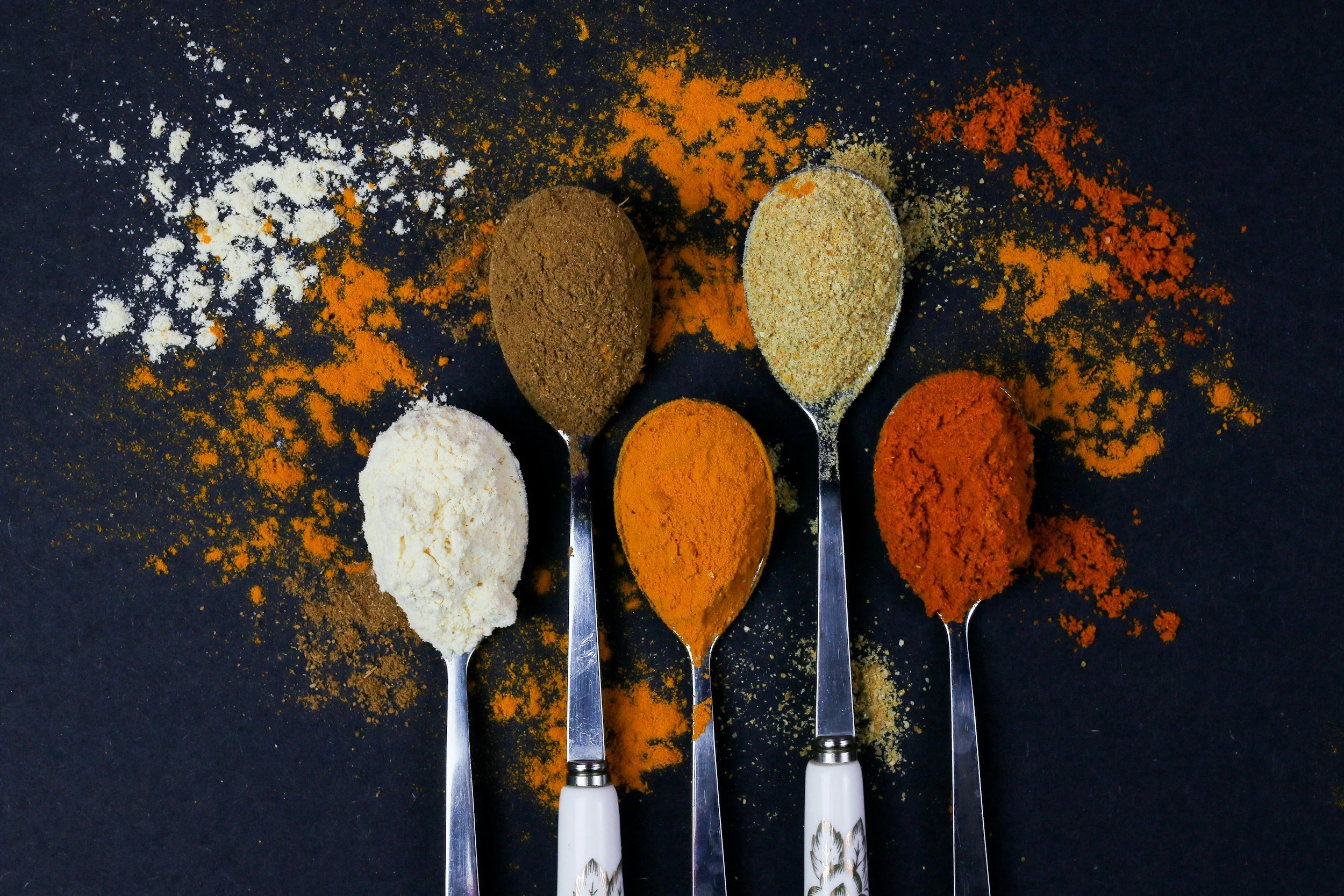 Five stainless steel spoons with white handles filled with different spices arranged on a black surface, with scattered spice powders around them. From left to right, the spices are white powder, brown powder, orange powder, beige powder, and red powder.