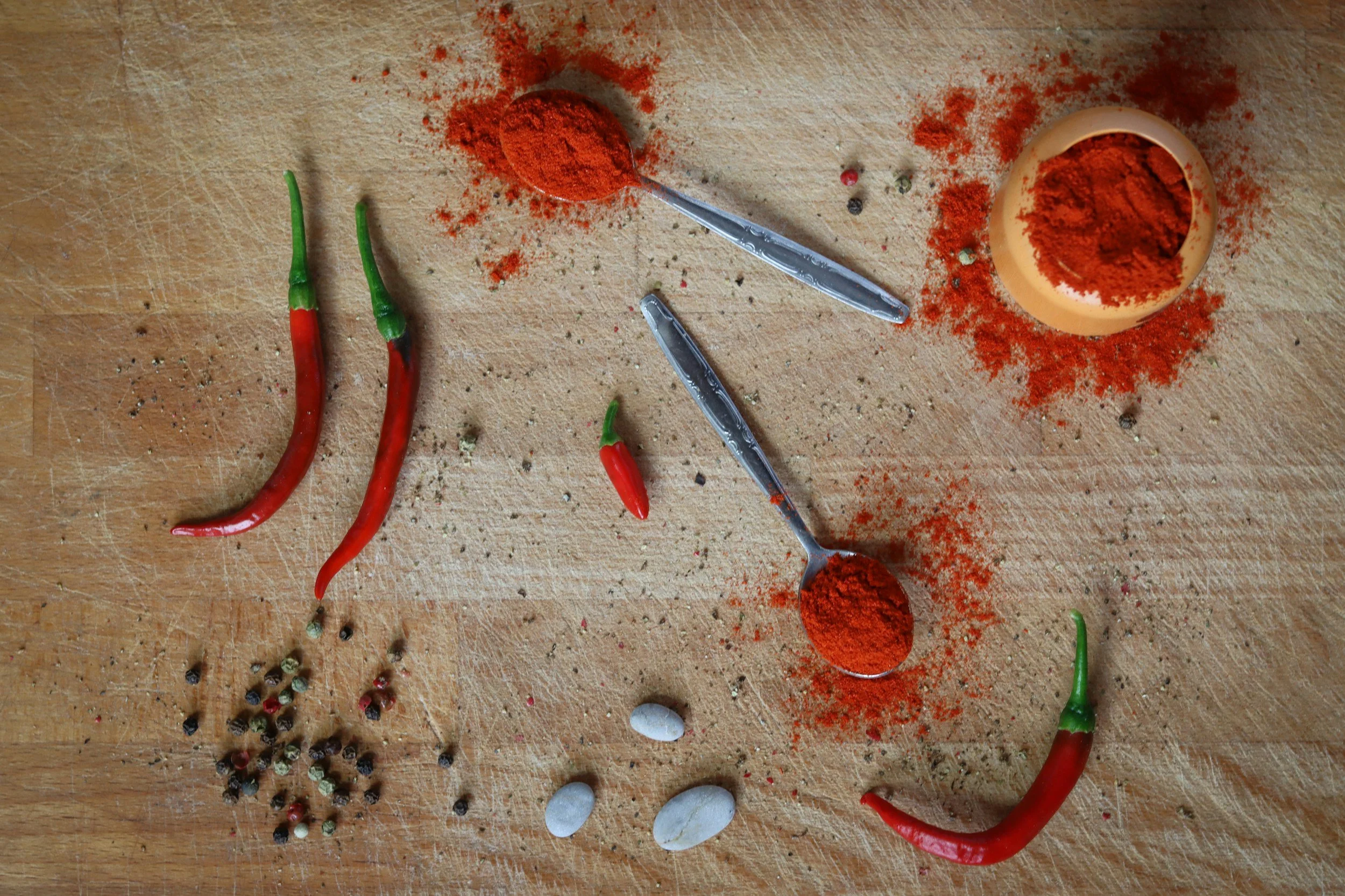 Red chili powder, chili peppers, peppercorns, and white stones on a wooden surface.