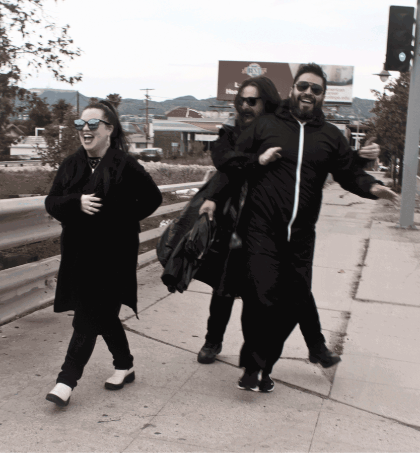 Three people laughing and walking on a sidewalk, two men carry a woman on their back, all wearing dark clothing and sunglasses, with hills and a billboard in the background.