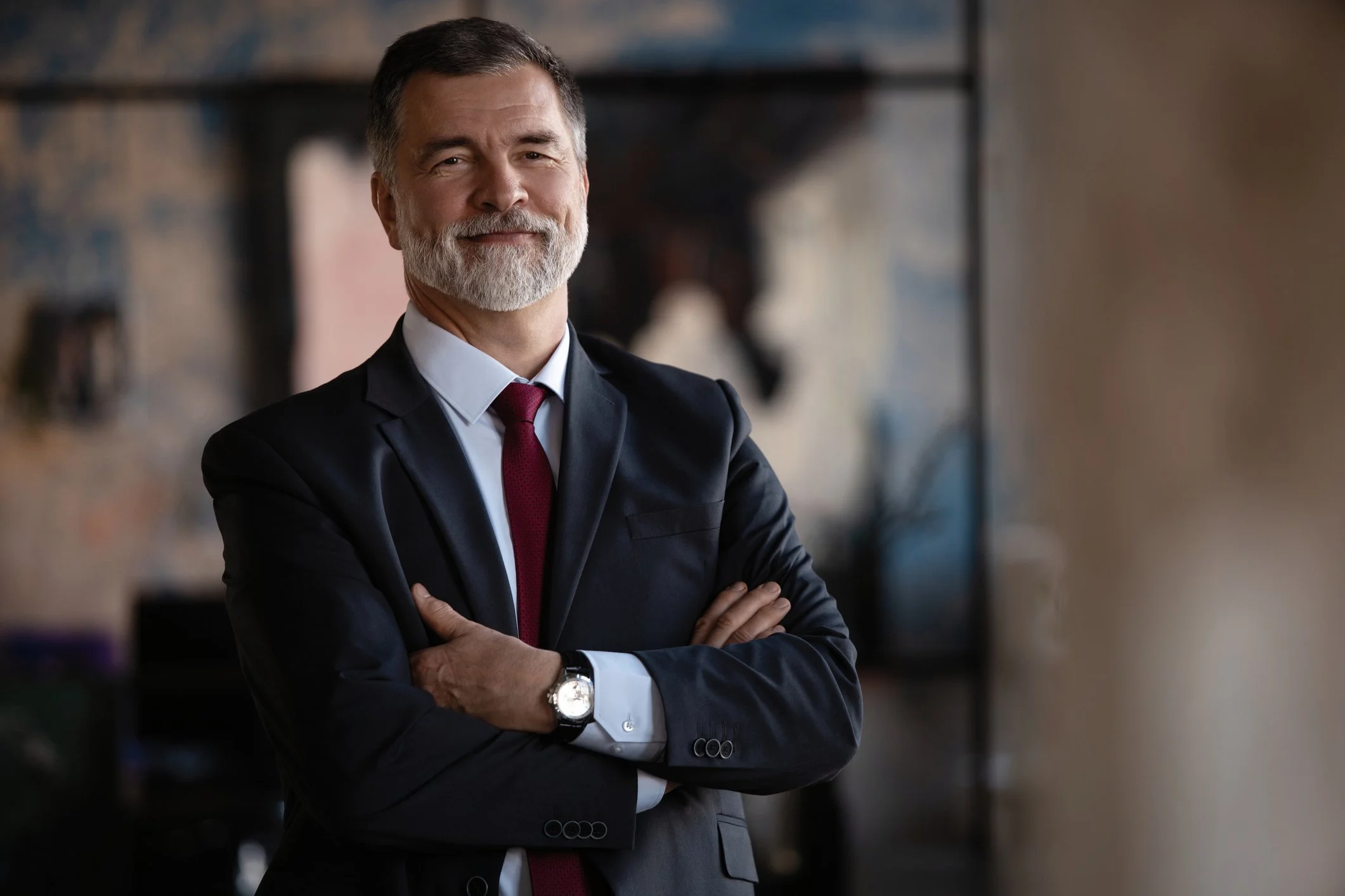A confident middle-aged man with gray hair and beard, dressed in a black suit, white shirt, and red tie, standing with arms crossed and smiling in an office setting.