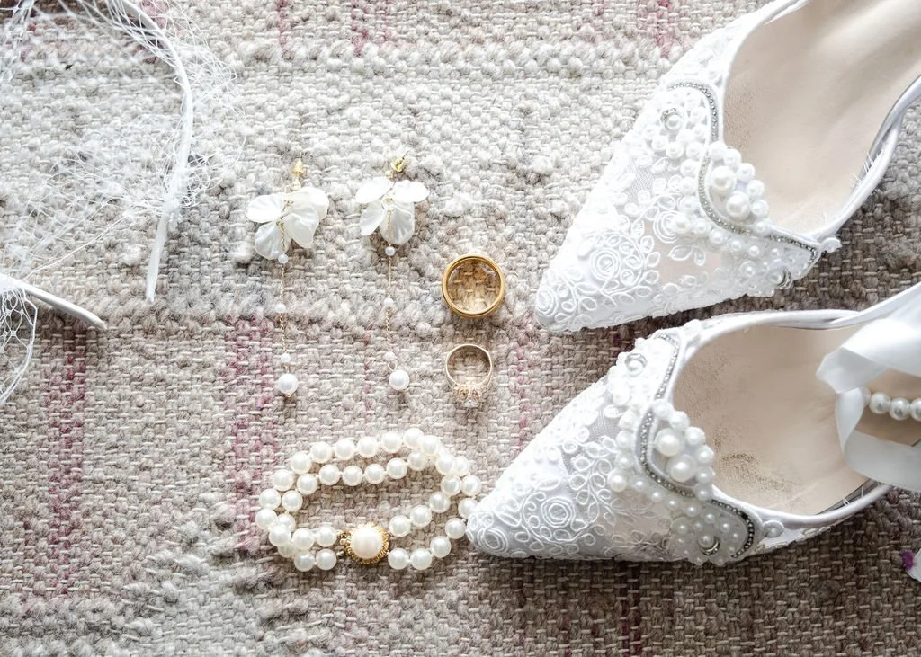 White bridal shoes with pearl and lace details, pearl jewelry including a bracelet and earrings, a gold ring, and a gold wedding band arranged on a textured beige and pink woven surface.
