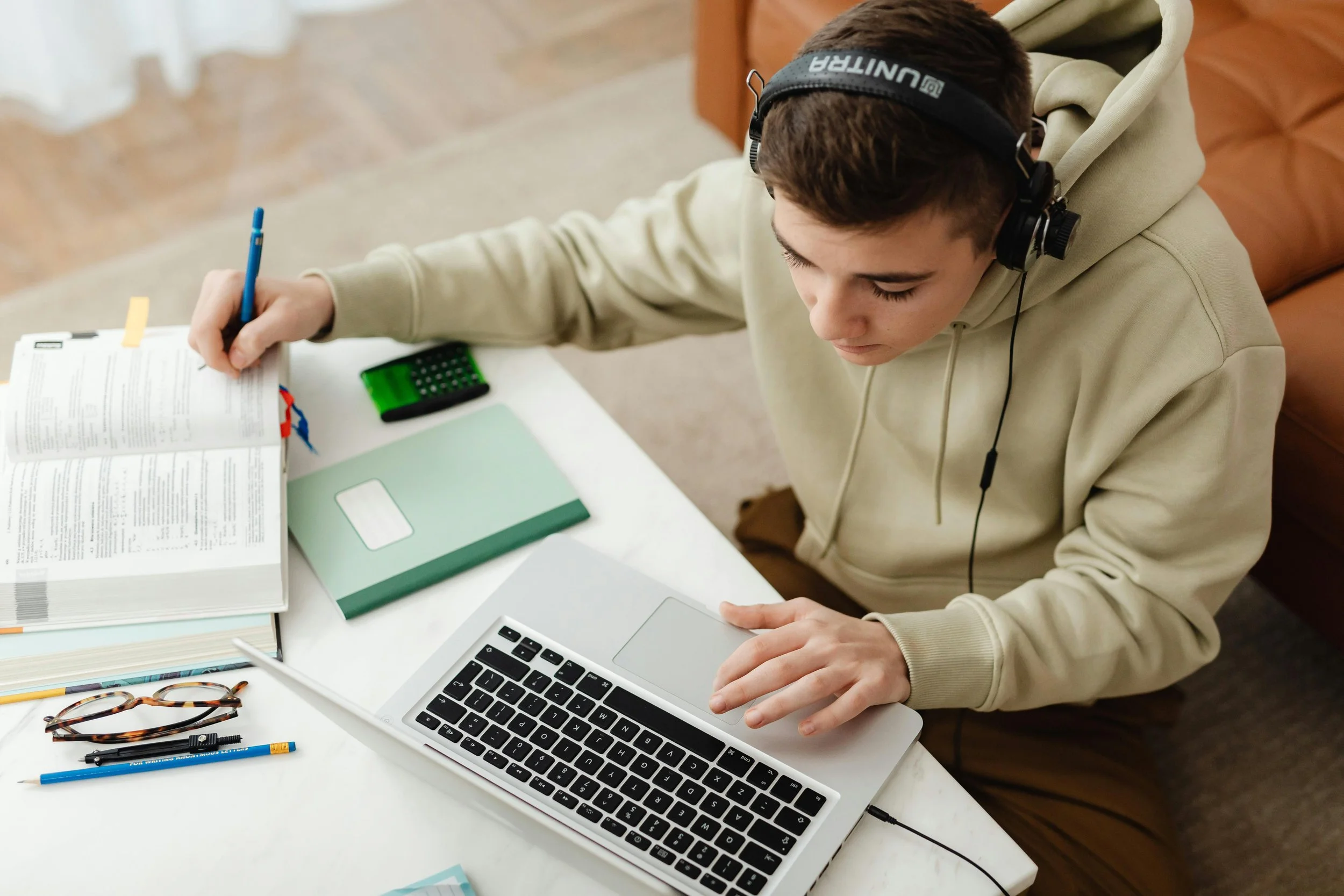 A young person wearing headphones sitting at a table, studying with a laptop, open book, notebook, calculator, eyeglasses, and pens.