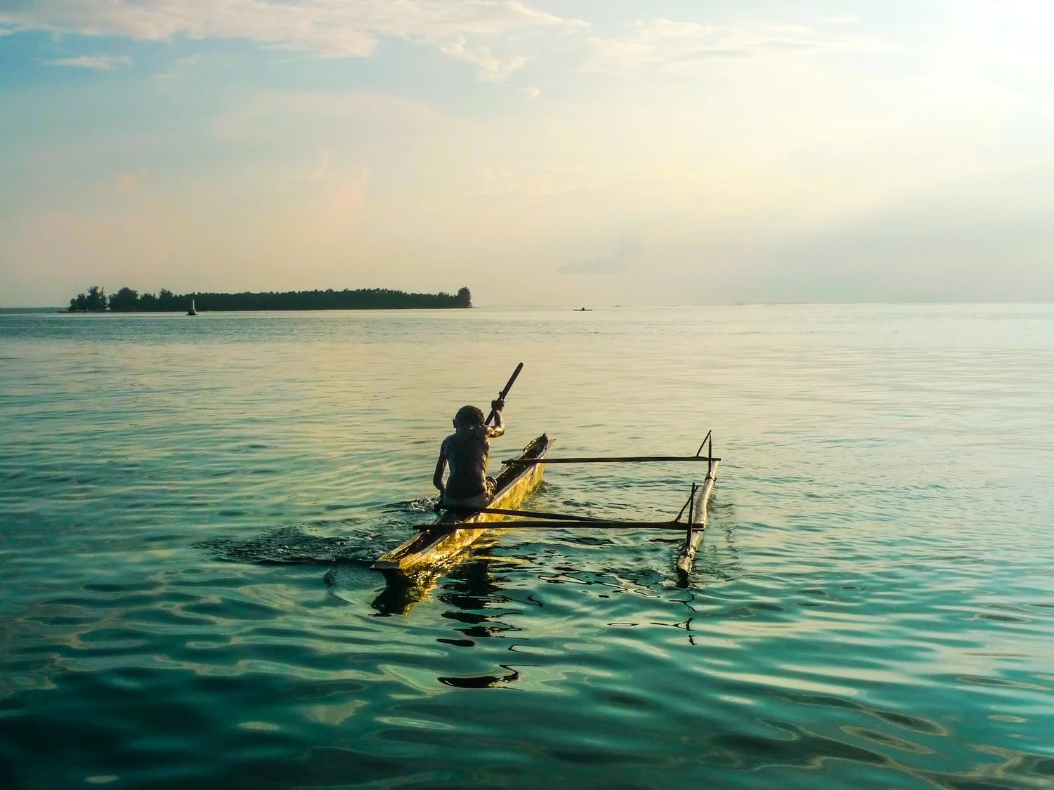 A person paddling a small, narrow boat with a wooden pole in calm water during sunset, with an island and other boats visible in the distance.