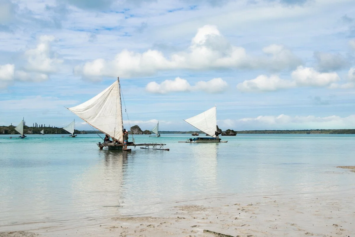 Sailboats with white sails floating on calm, turquoise water near sandy beach under partly cloudy sky.