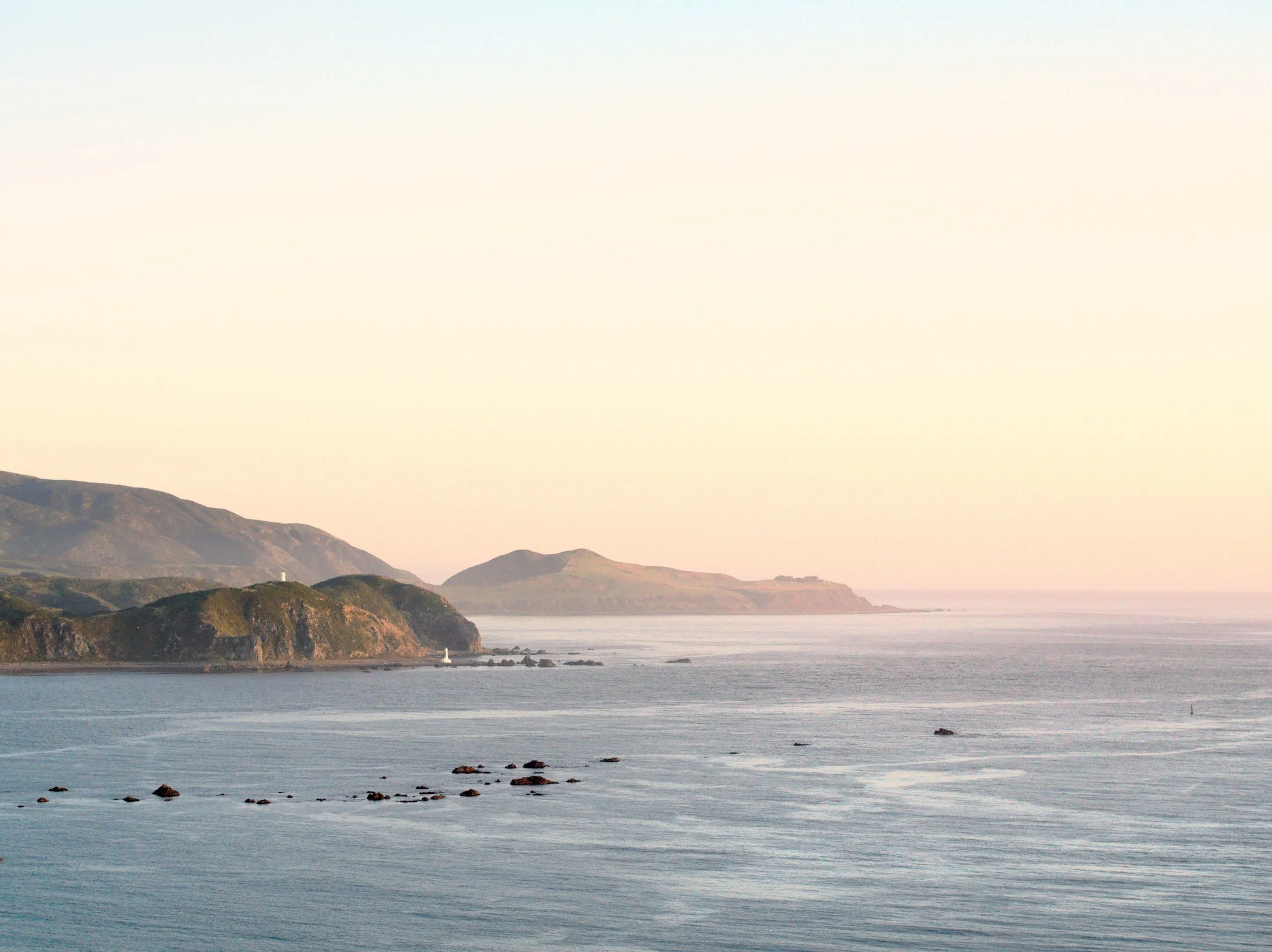 A coastal landscape with ocean water in the foreground, rocky shoreline, and hills or mountains in the background under a clear or slightly hazy sky.