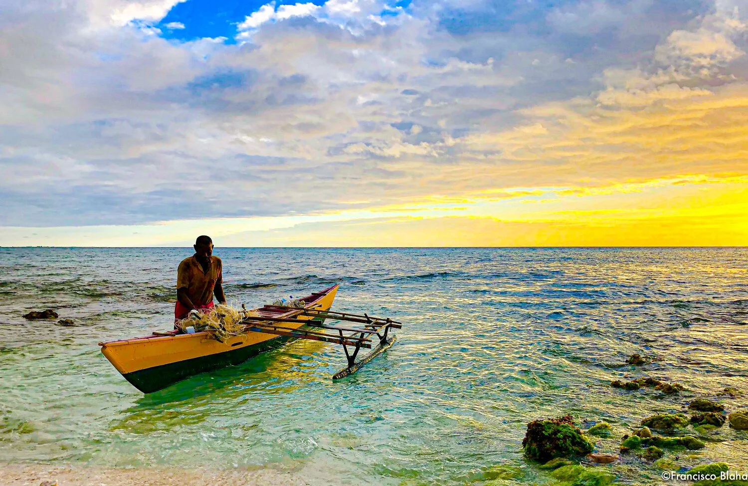 A man on a yellow boat near the shore at sunset or sunrise, with rocks and seaweed on the beach in the foreground and a colorful sky with clouds.